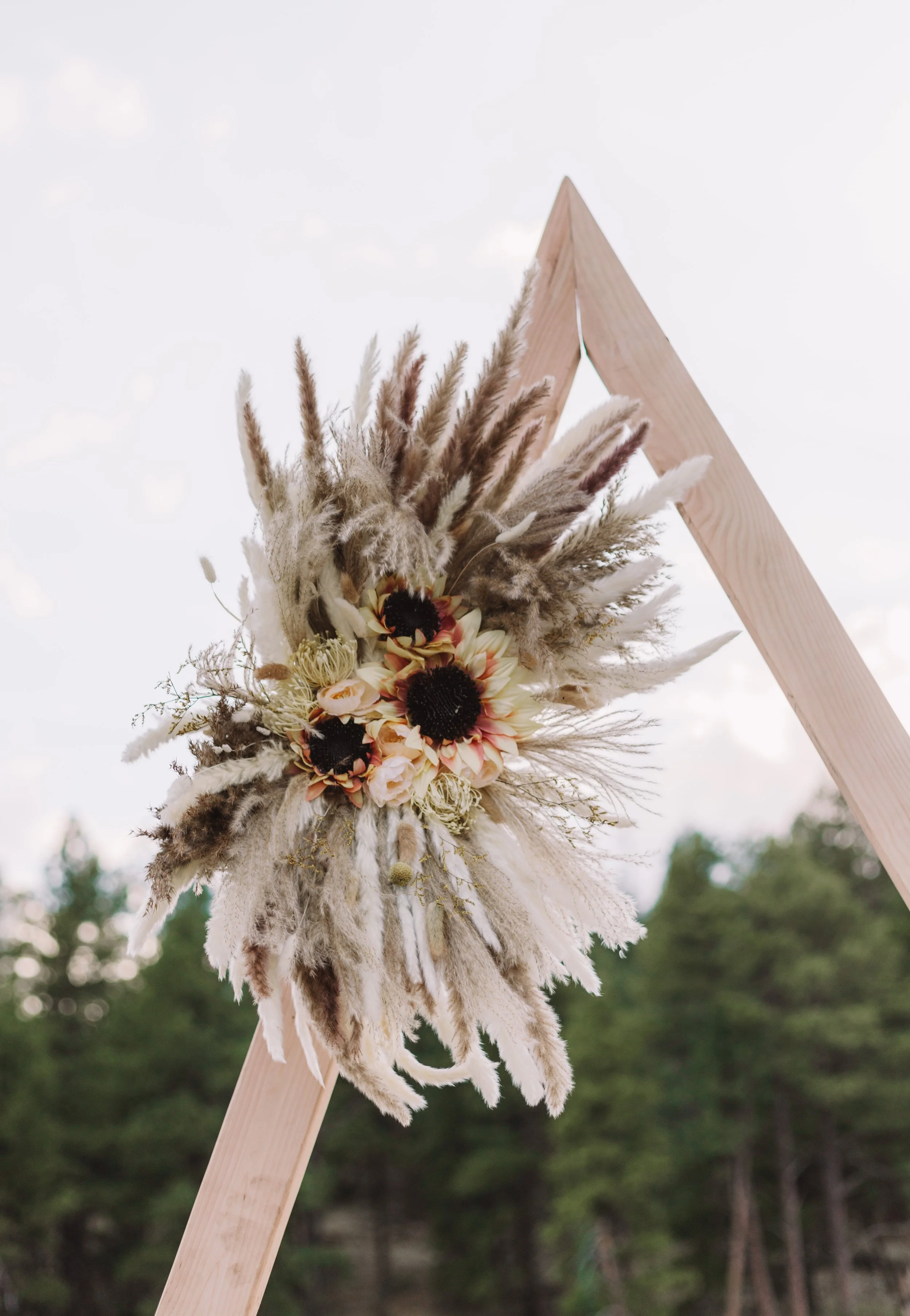 Dried floral arrangement with sunflowers and pampas grass attached to a wooden triangular wedding arch outdoors.