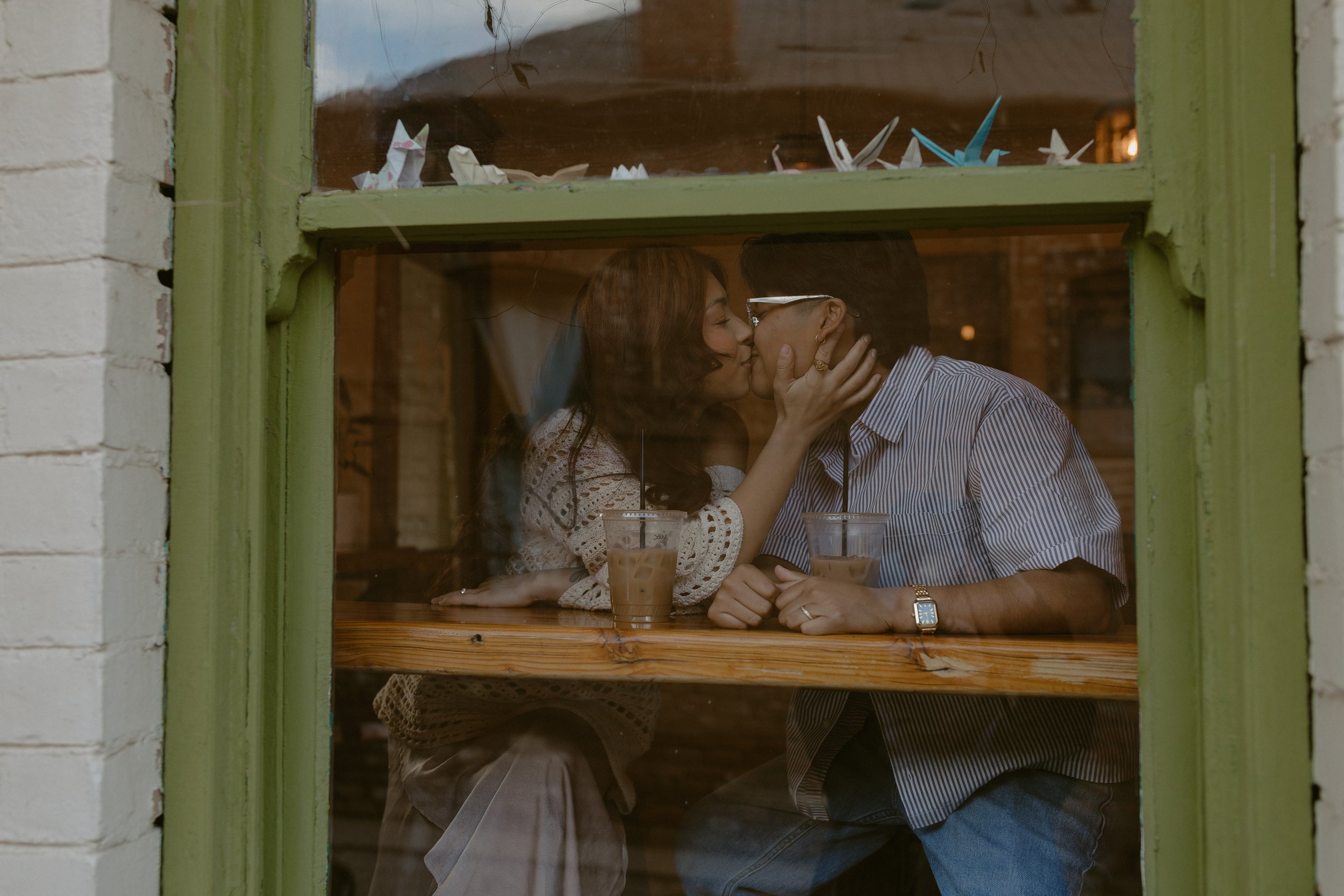 A couple sharing a kiss inside a cafe, seen through a window decorated with origami animals.