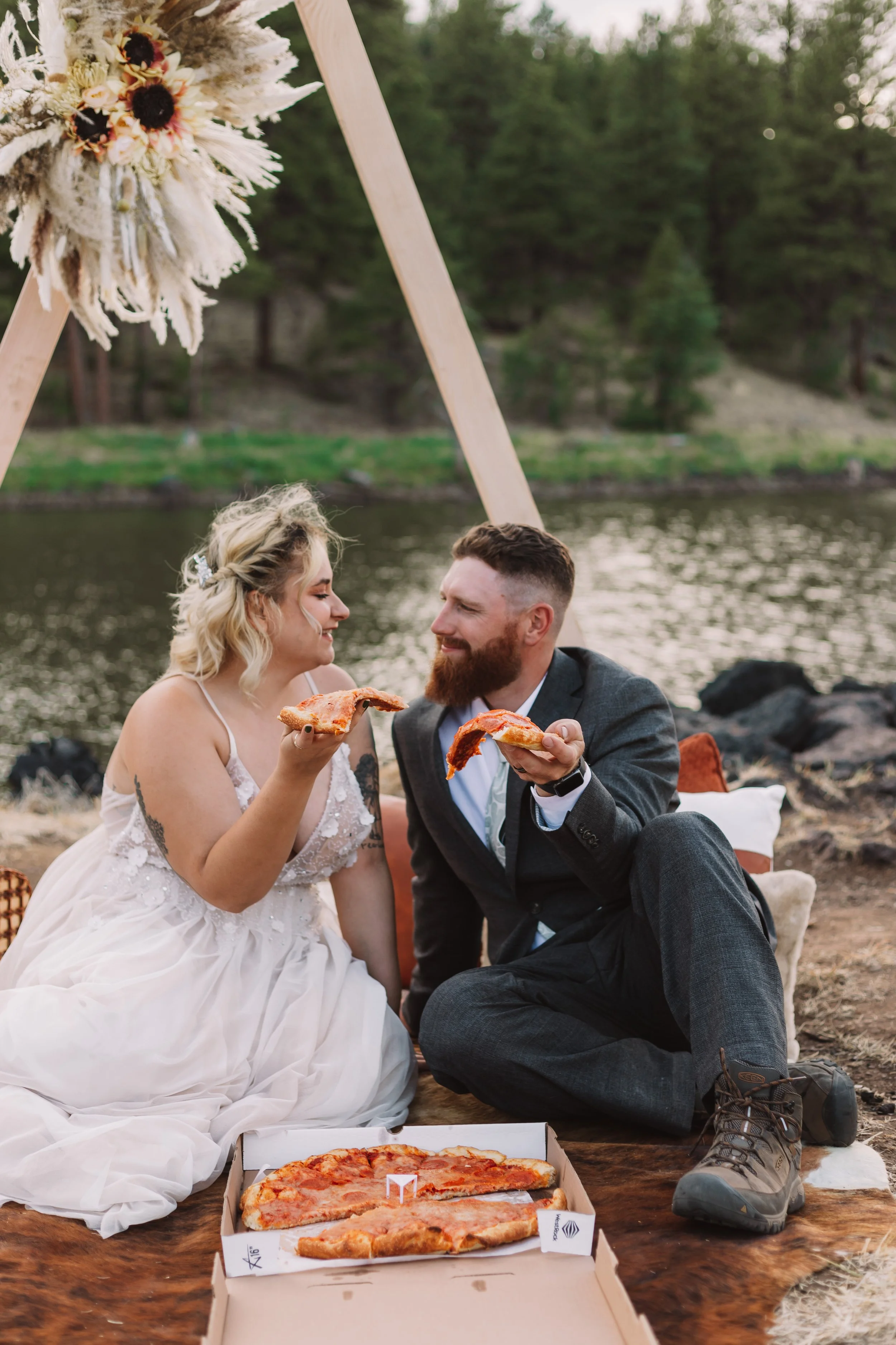 A couple in formal attire sitting by a lakeside, sharing pizza. The woman is in a wedding dress, and the man in a suit. They are seated on a rug, with a wooden arch decorated with flowers in the background. Trees and water are visible.