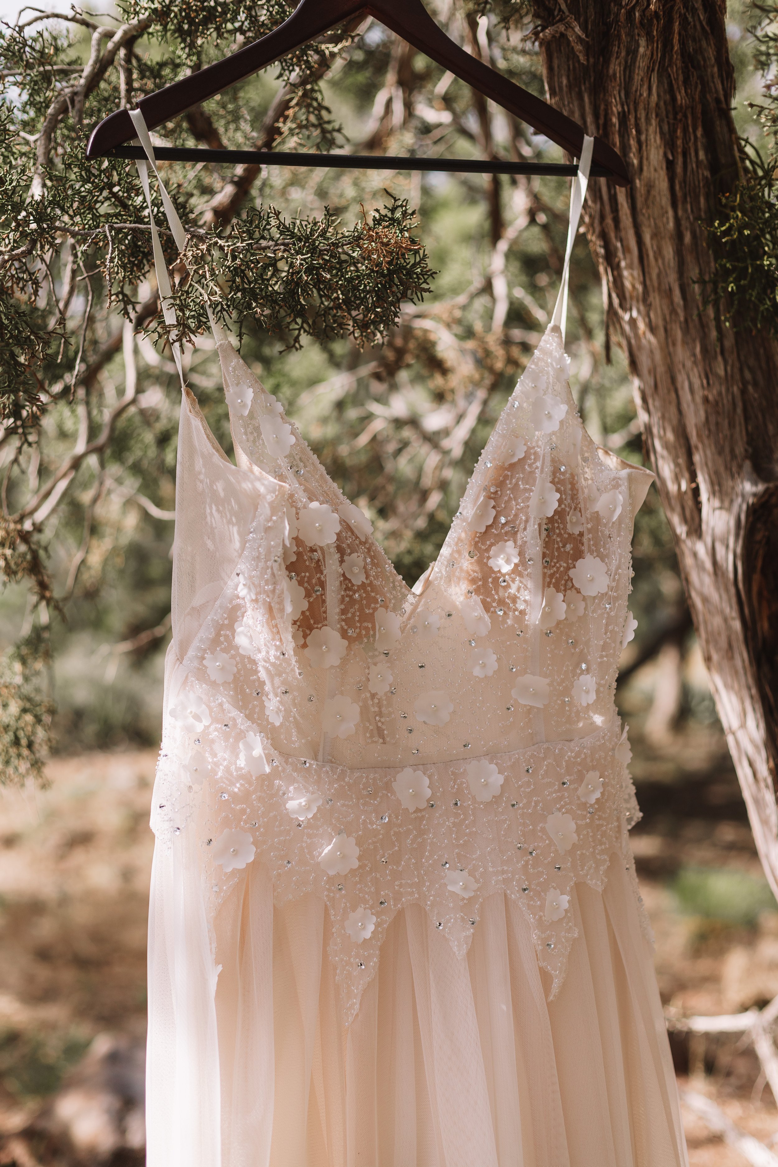 A white and floral wedding dress on a wooden hanger outdoors, with a backdrop of trees.