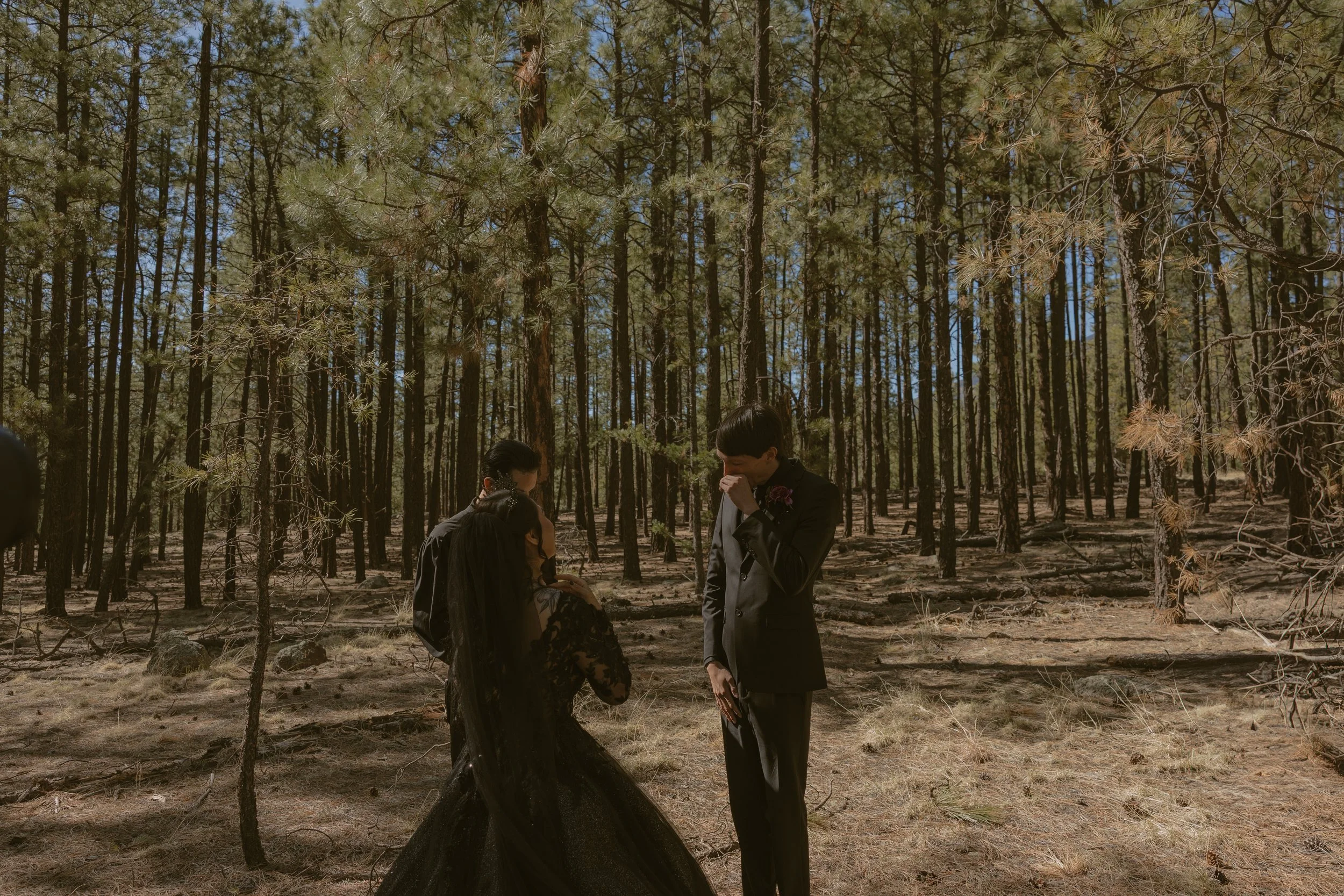 A couple dressed in formal attire standing in a forest with tall trees, one woman in a black dress and one man in a suit, both covering their faces with their hands.