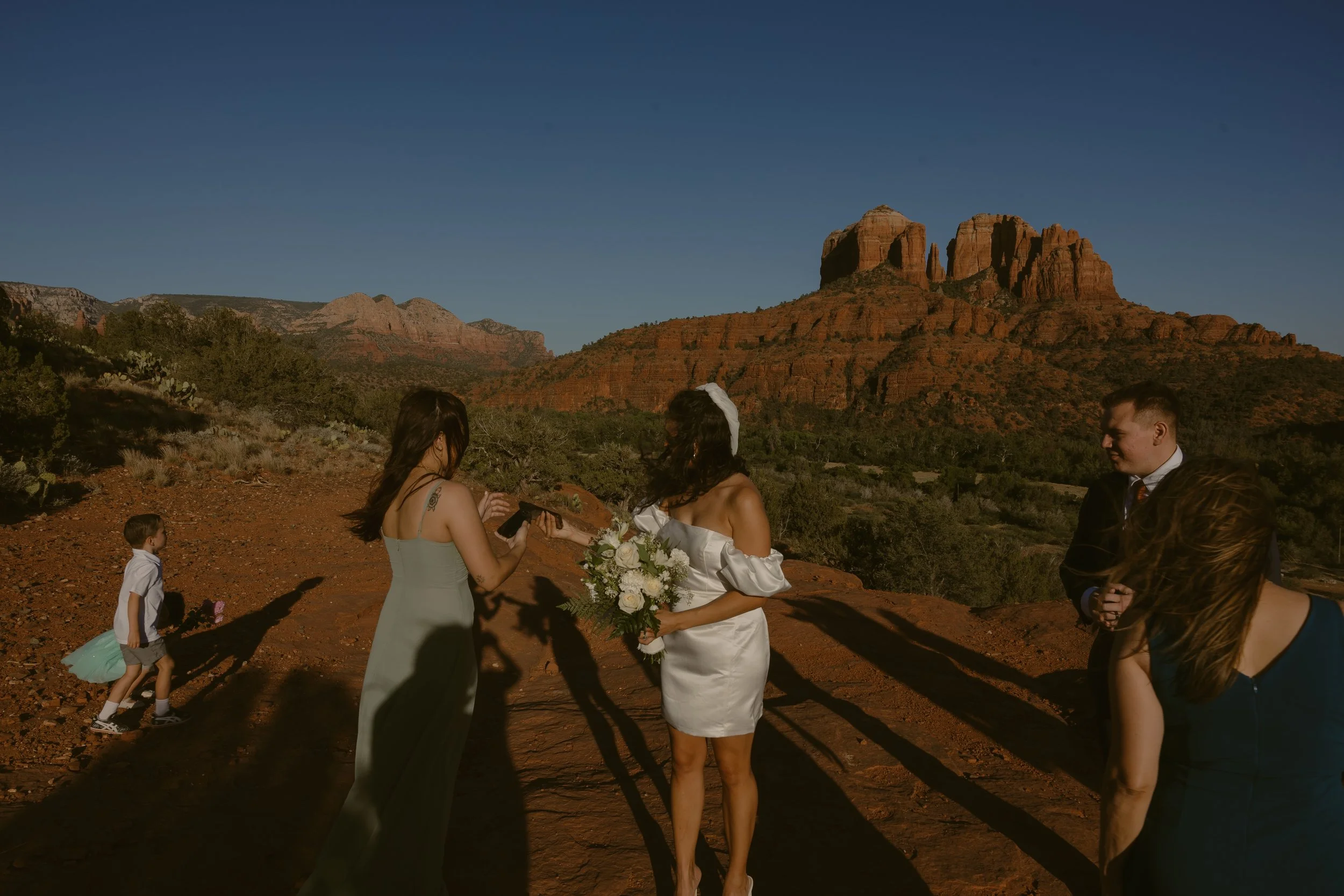 A wedding ceremony taking place outdoors with red rock formations in the background. The bride is holding a bouquet, and others are standing around her.