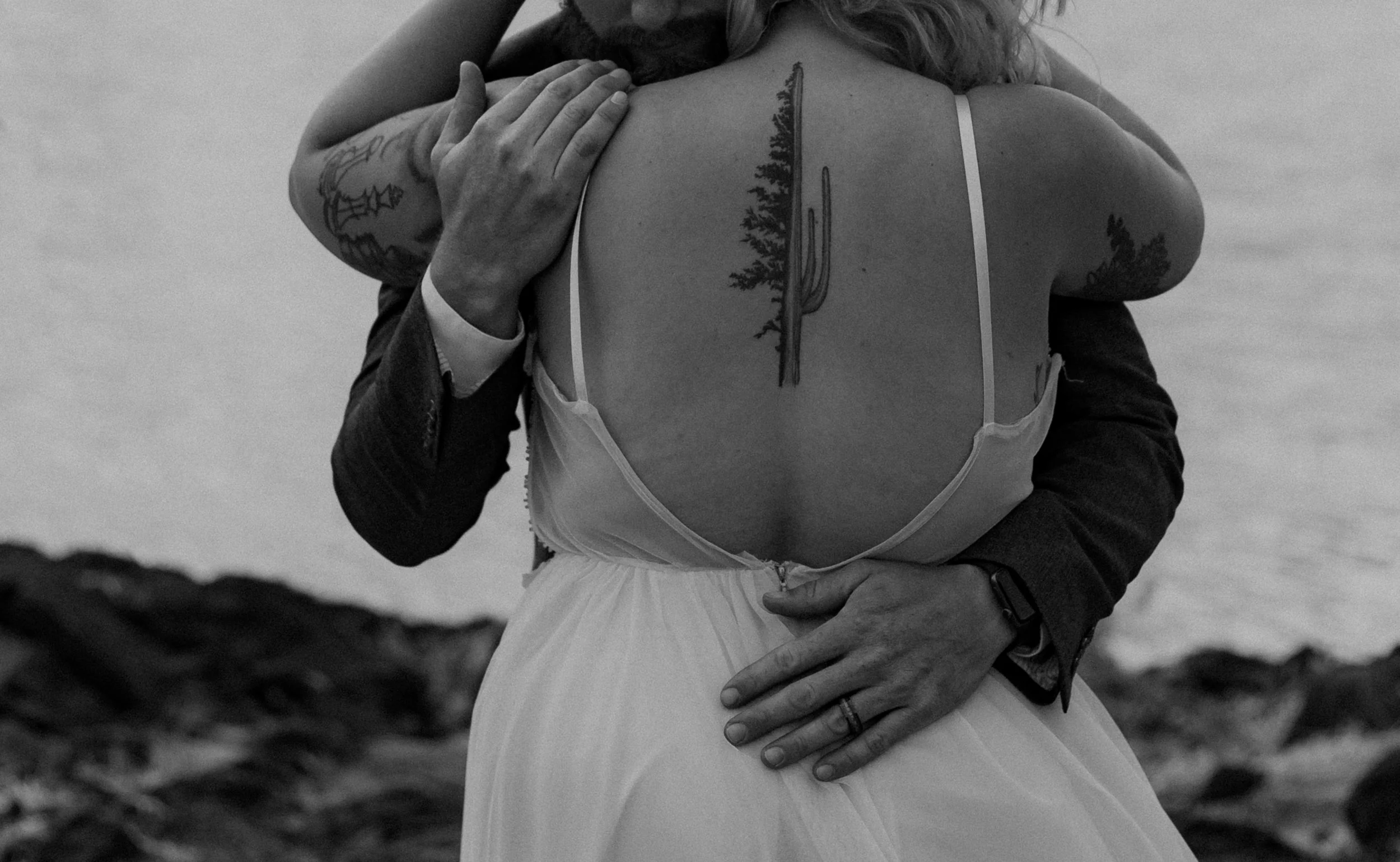 Black and white photo of a couple embracing, with the focus on the woman's back showing a tattoo of a cactus. She is wearing a backless dress, and the man's arms are around her.