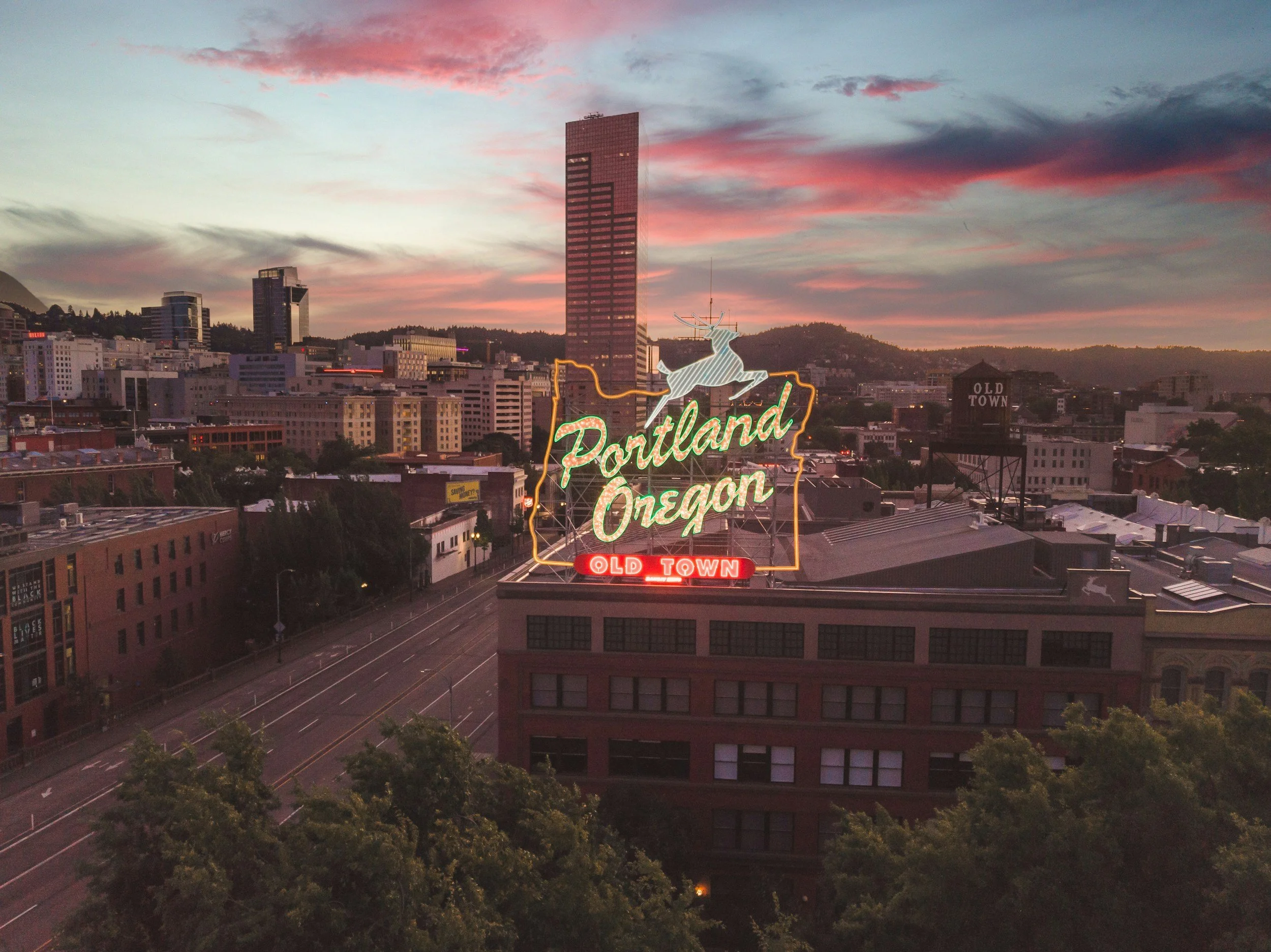 City skyline during sunset with a neon sign reading 'Portland Oregon' featuring a reindeer illustration, and old town water tower in the background.