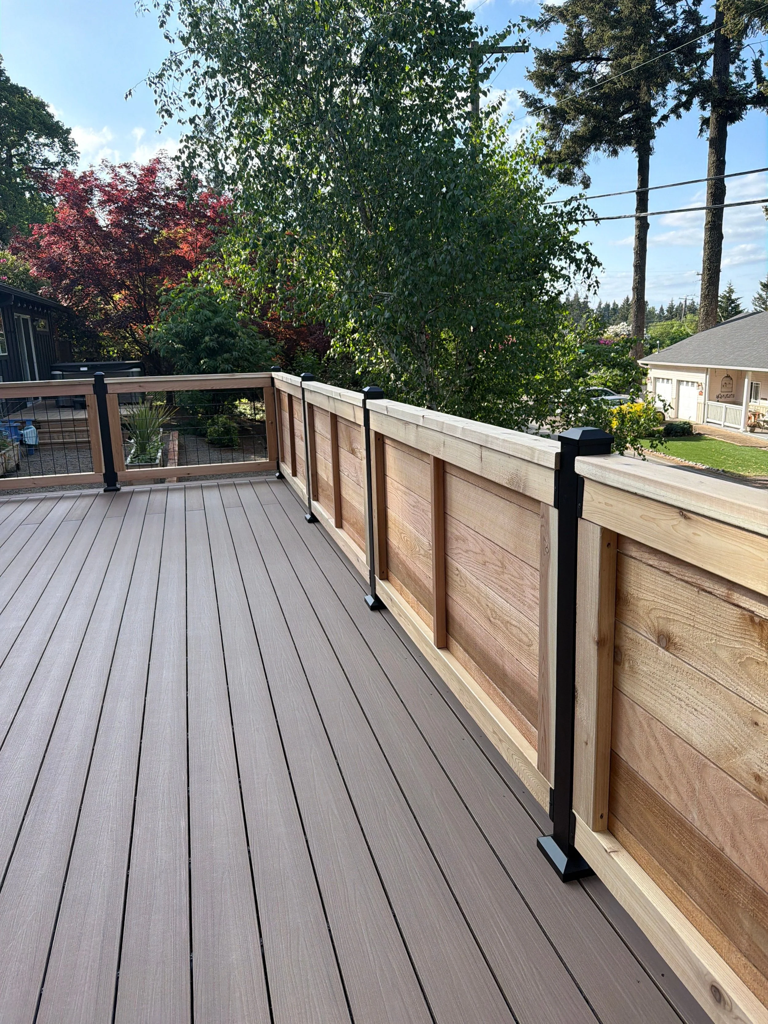 View of a backyard deck with a wooden railing, overlooking trees, neighboring houses, and a partly cloudy sky.