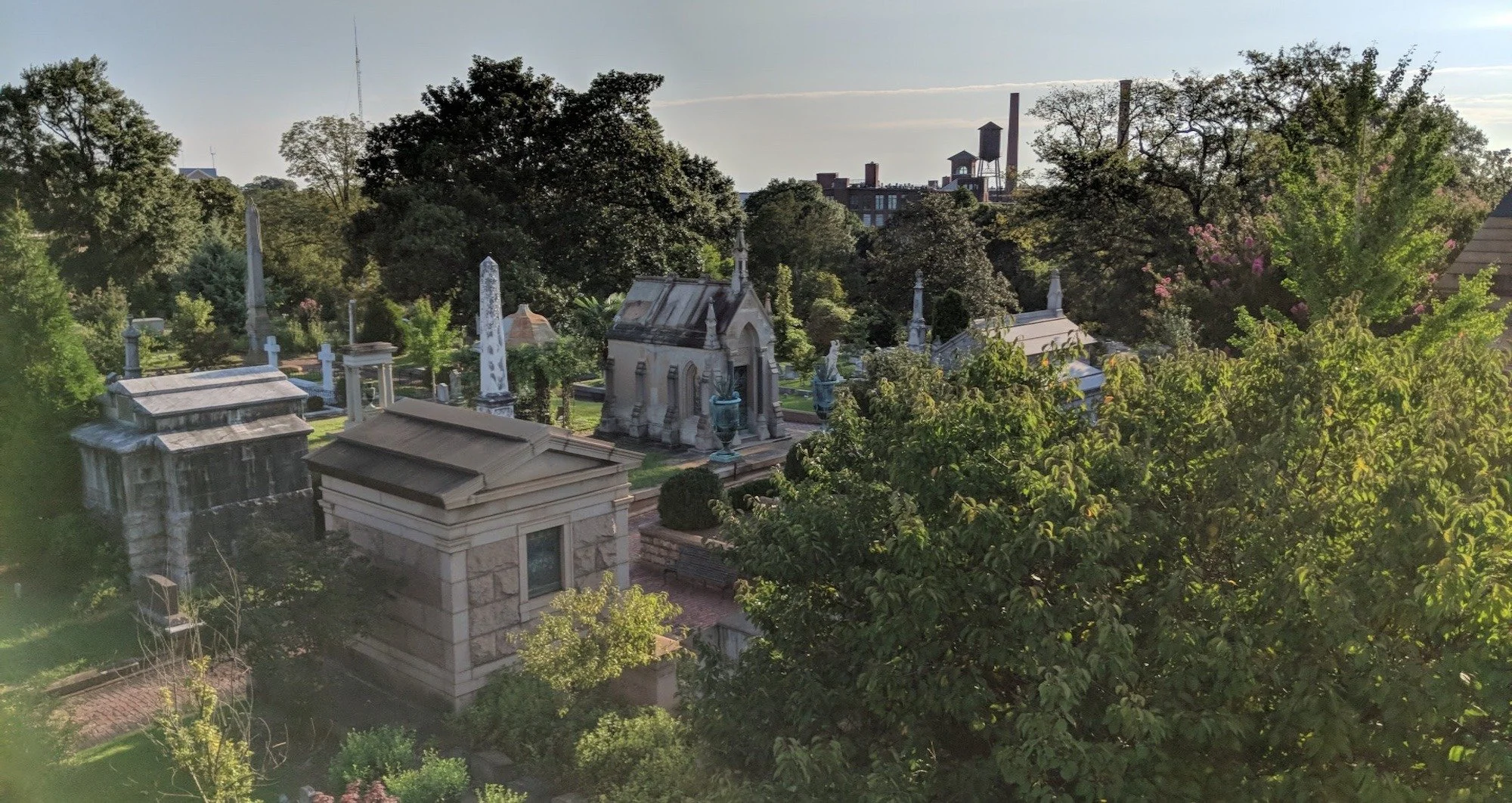 A graveyard with tall trees and old stone mausoleums, some with statues, under a partly cloudy sky with a city skyline and industrial buildings in the background.