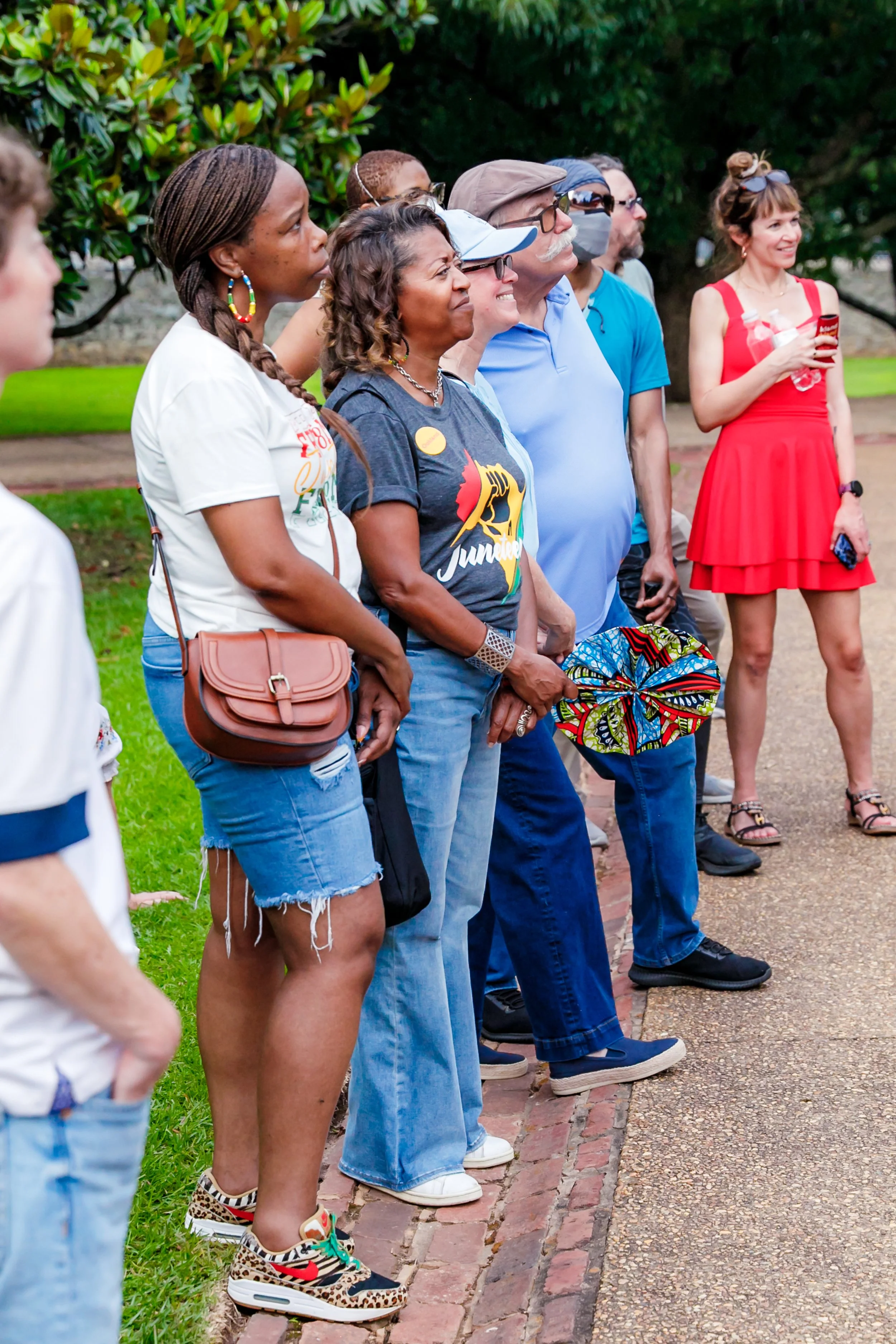 Group of diverse people standing outdoors on a brick path, attentively listening during a gathering or event, with trees and greenery in the background.