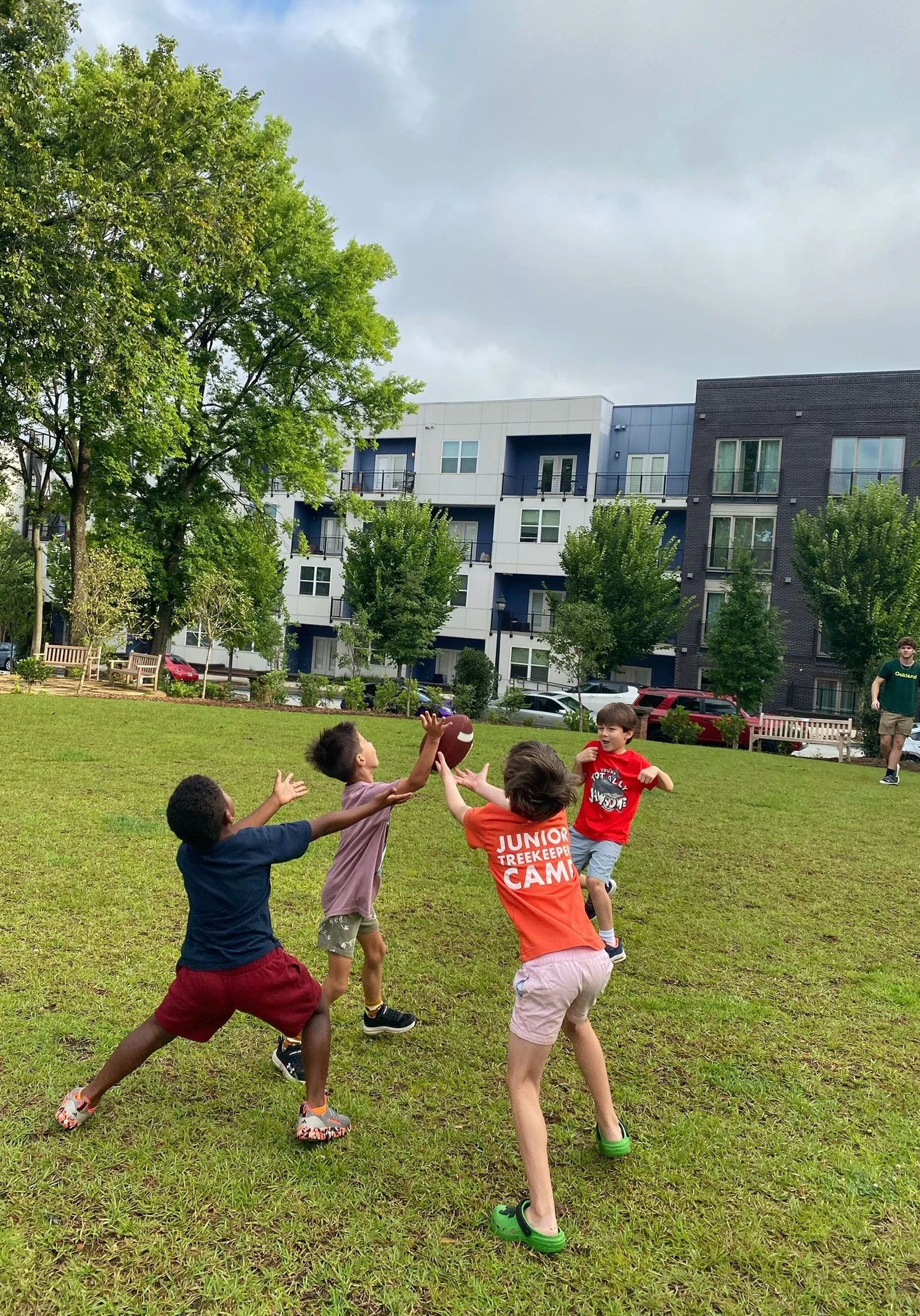 Children playing catch with a football in a grassy park area, with trees and modern apartment buildings in the background on a cloudy day.