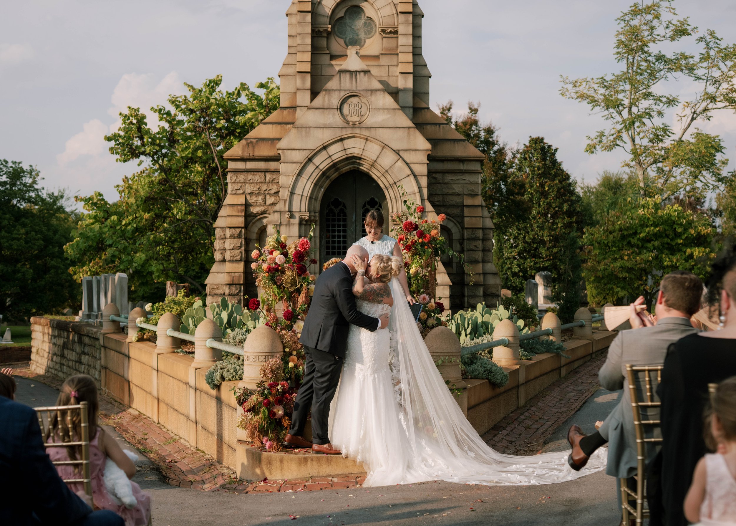 A wedding ceremony outdoors near a stone church with floral arrangements, where a couple is exchanging vows with an officiant. Guests are seated nearby, and the scene is set in a lush green environment.