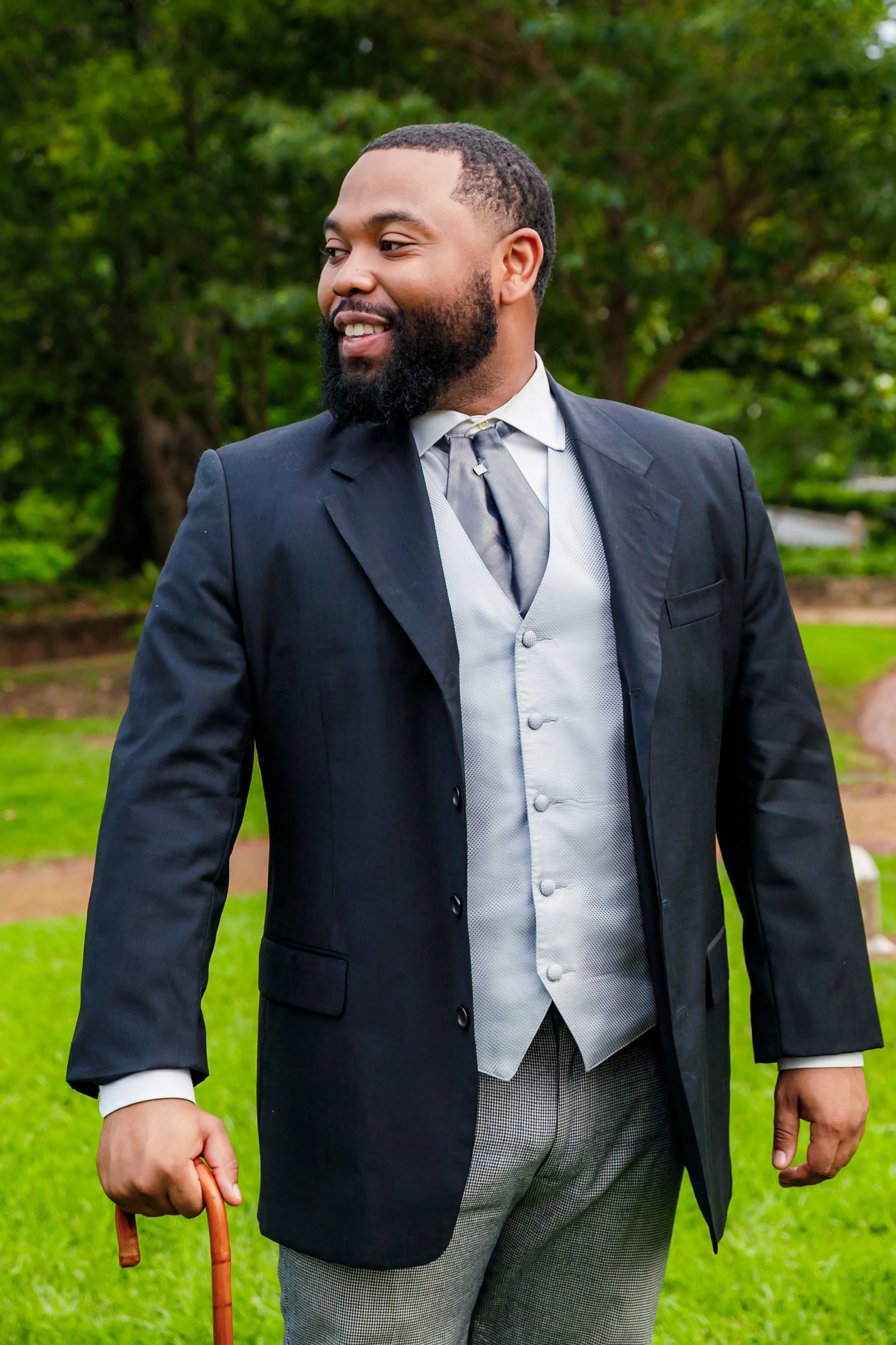 A man wearing a formal suit, gray vest, and tie, holding a walking cane, standing outdoors with greenery in the background.