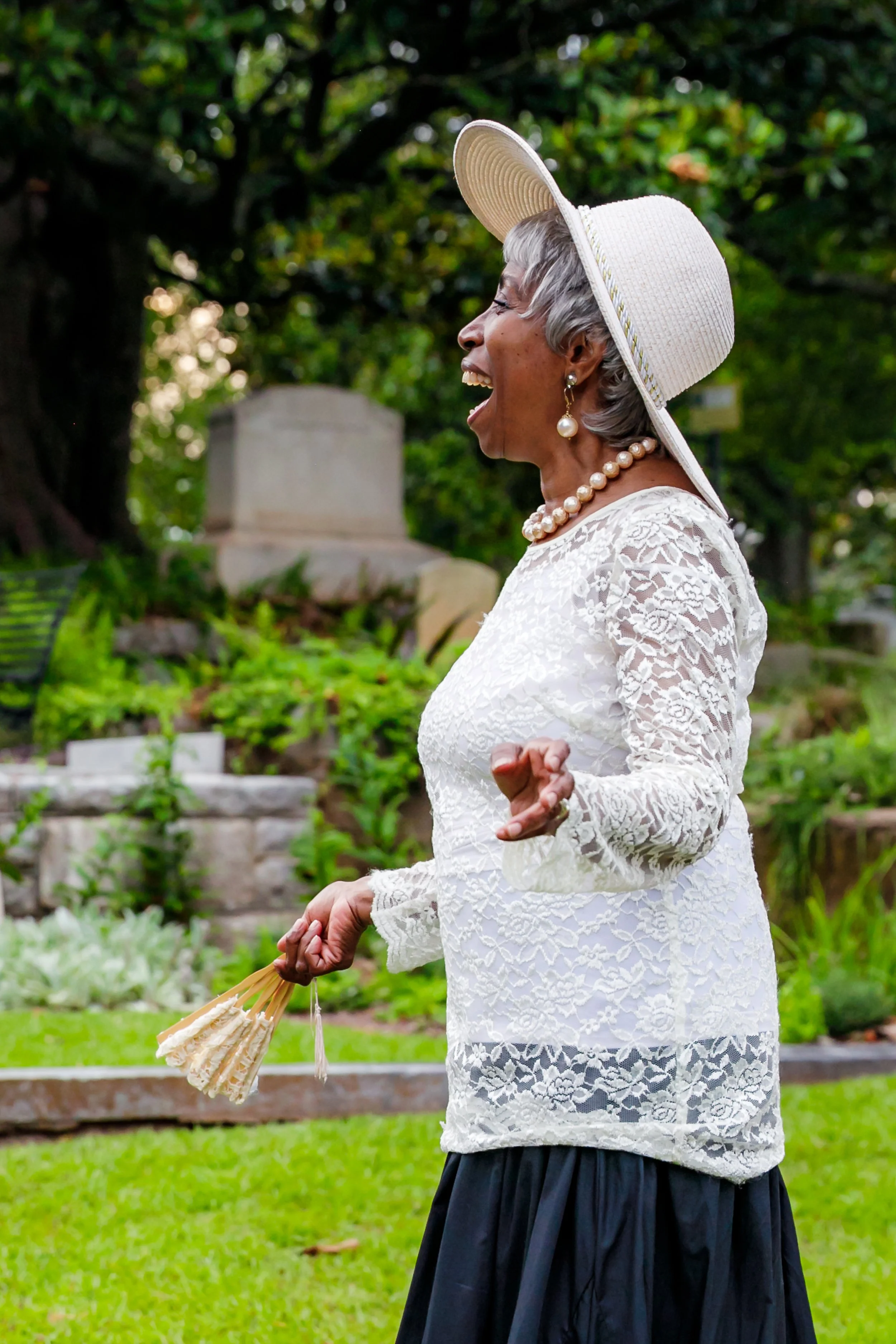 An elderly woman with gray hair wearing a large floppy sun hat, pearl earrings, and a pearl necklace, dressed in a white lace top and black skirt, laughing outdoors in a garden with lush greenery and a stone monument in the background.