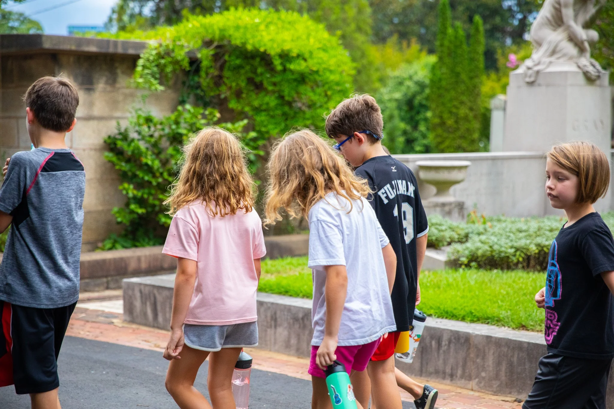 A group of children walking outdoors in a park-like setting with greenery and a stone monument or sculpture in the background.