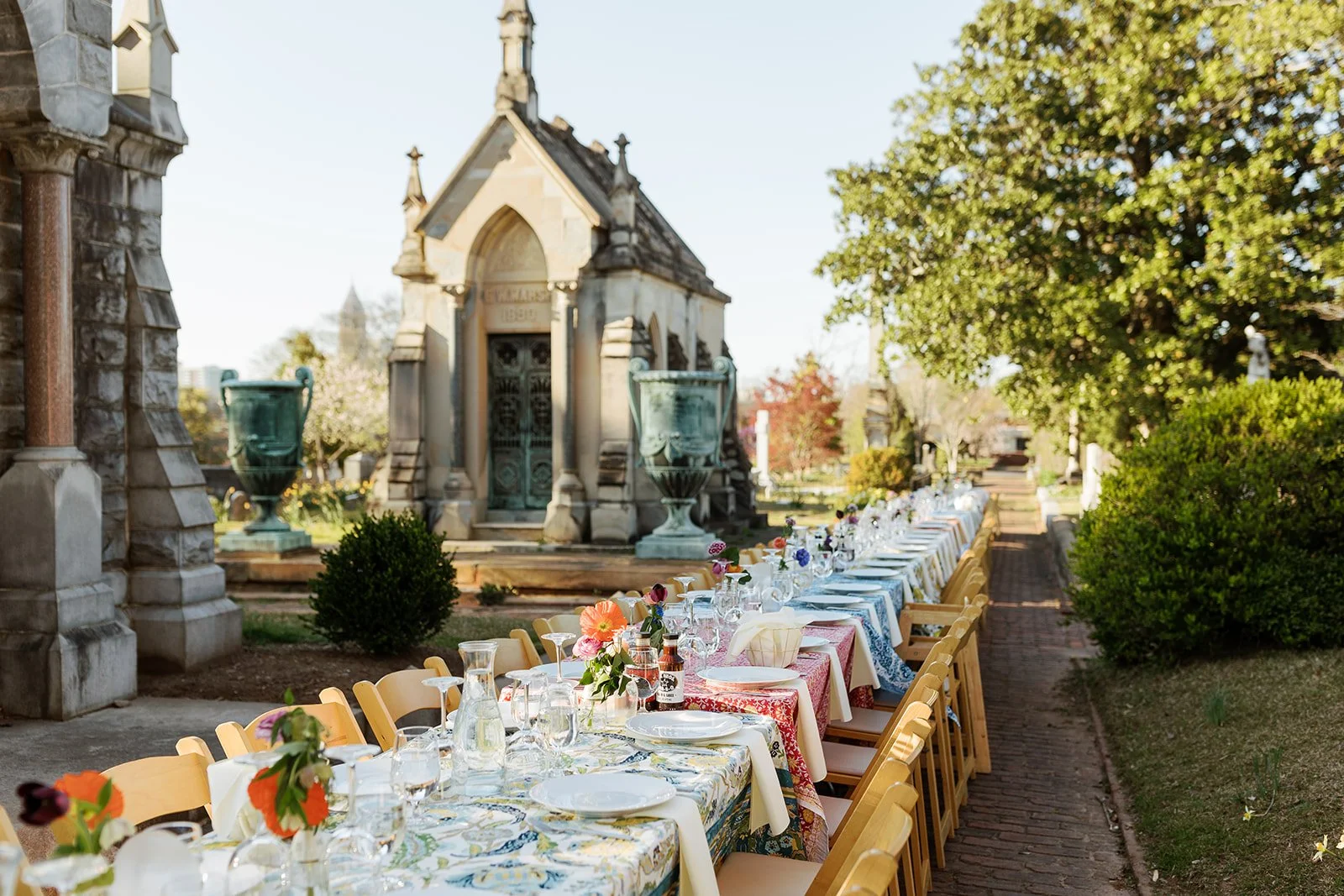 Long outdoor dining table set with colorful tablecloths, floral arrangements, glasses, and white plates in a garden with historic tombstones and monuments, surrounded by trees and greenery.