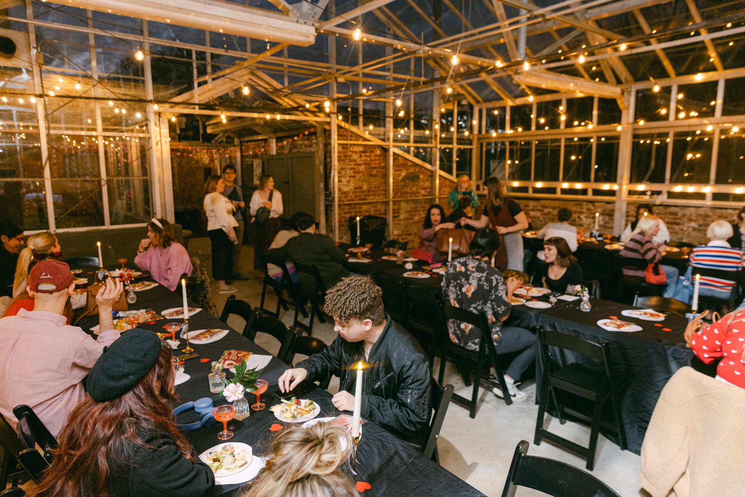 A lively indoor gathering with guests seated at black tables decorated with candles and small floral centerpieces, under warm string lights in a partly open brick-walled space.