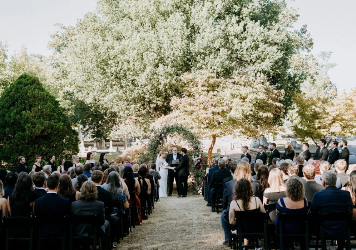 A wedding ceremony outdoors in front of a large blooming tree, with guests seated in rows and the couple exchanging vows under an arch decorated with flowers.