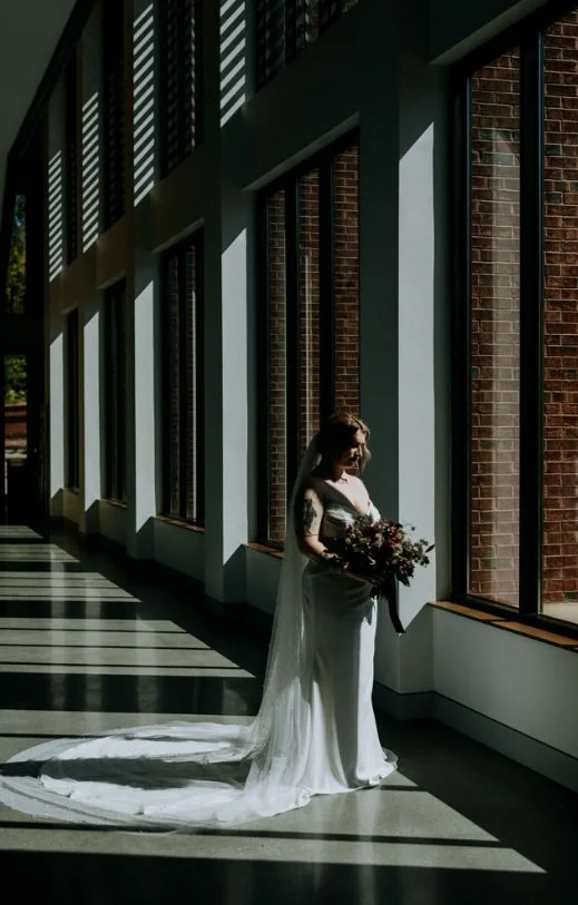 A bride with a bouquet standing inside near large windows, sunlight casting shadows on the floor and walls.