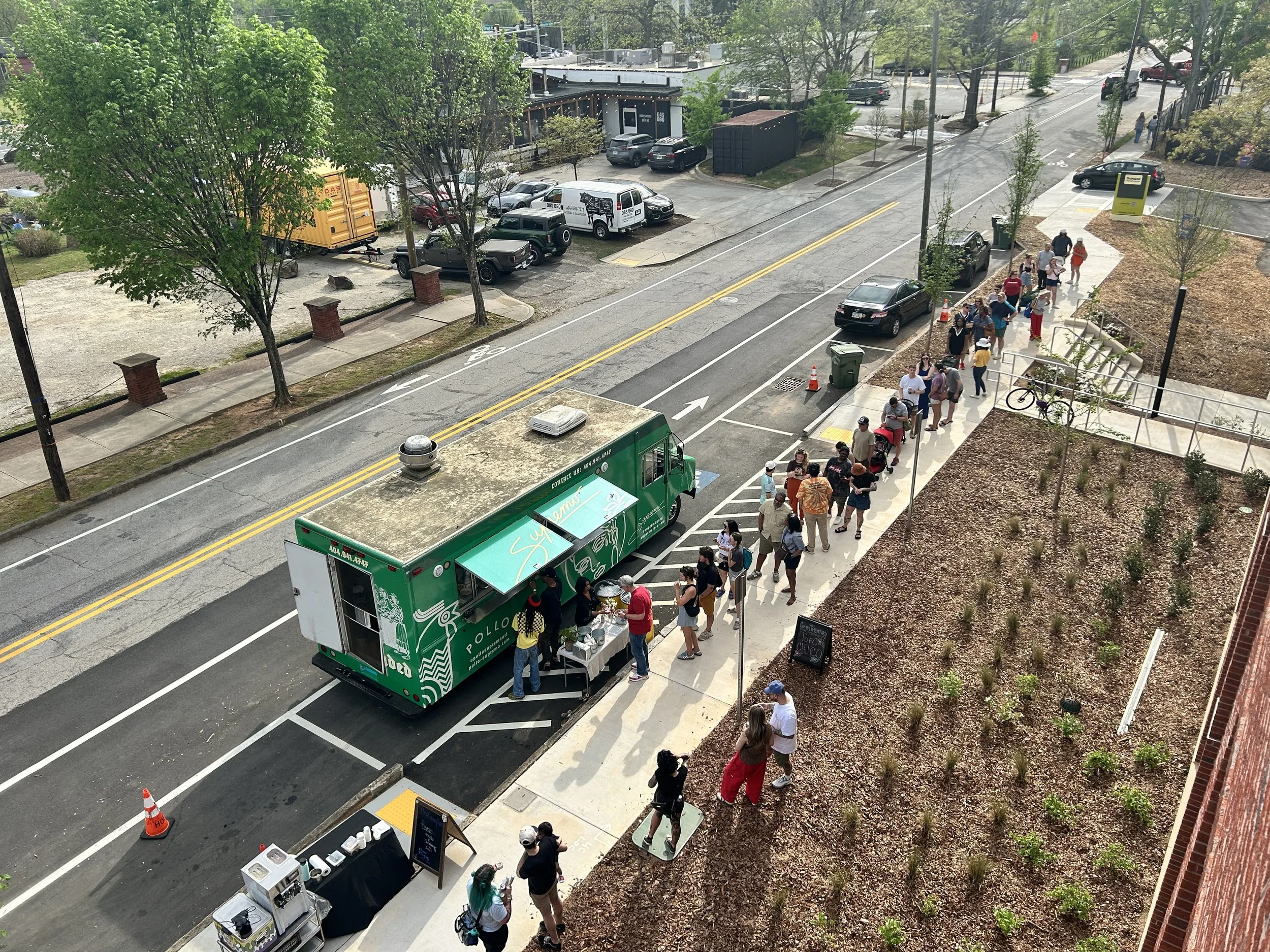 A food truck with a green design and signage serving customers in a line on a city street, with people waiting and ordering, some carrying food and beverages, and a sidewalk with small plants and a chalkboard sign.