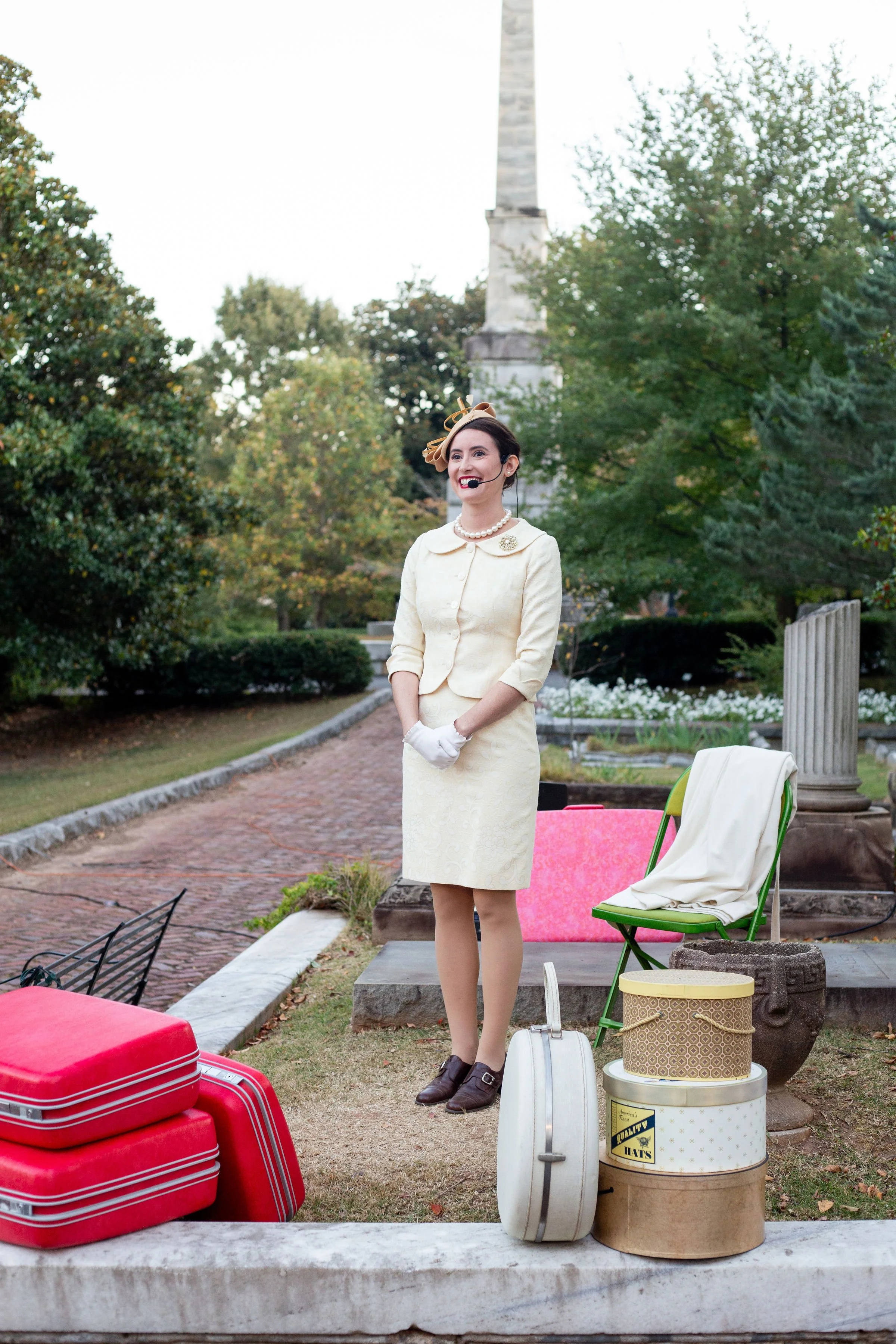 A woman dressed in vintage clothing, wearing a pale yellow dress with a matching jacket, black shoes, and a necklace, stands outdoors with a headset microphone. She is smiling and appears to be speaking or performing. Around her are colorful suitcases, a white bag, and a hat box, with lush green trees and a monument in the background.
