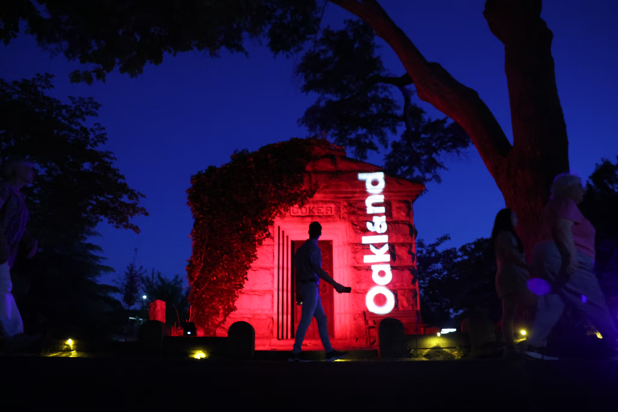 A nighttime scene with a historic monument illuminated in red and white lights. The monument features the word "COKE" prominently displayed on its side. Several people are walking or standing nearby, and trees frame the scene against a dark blue sky.