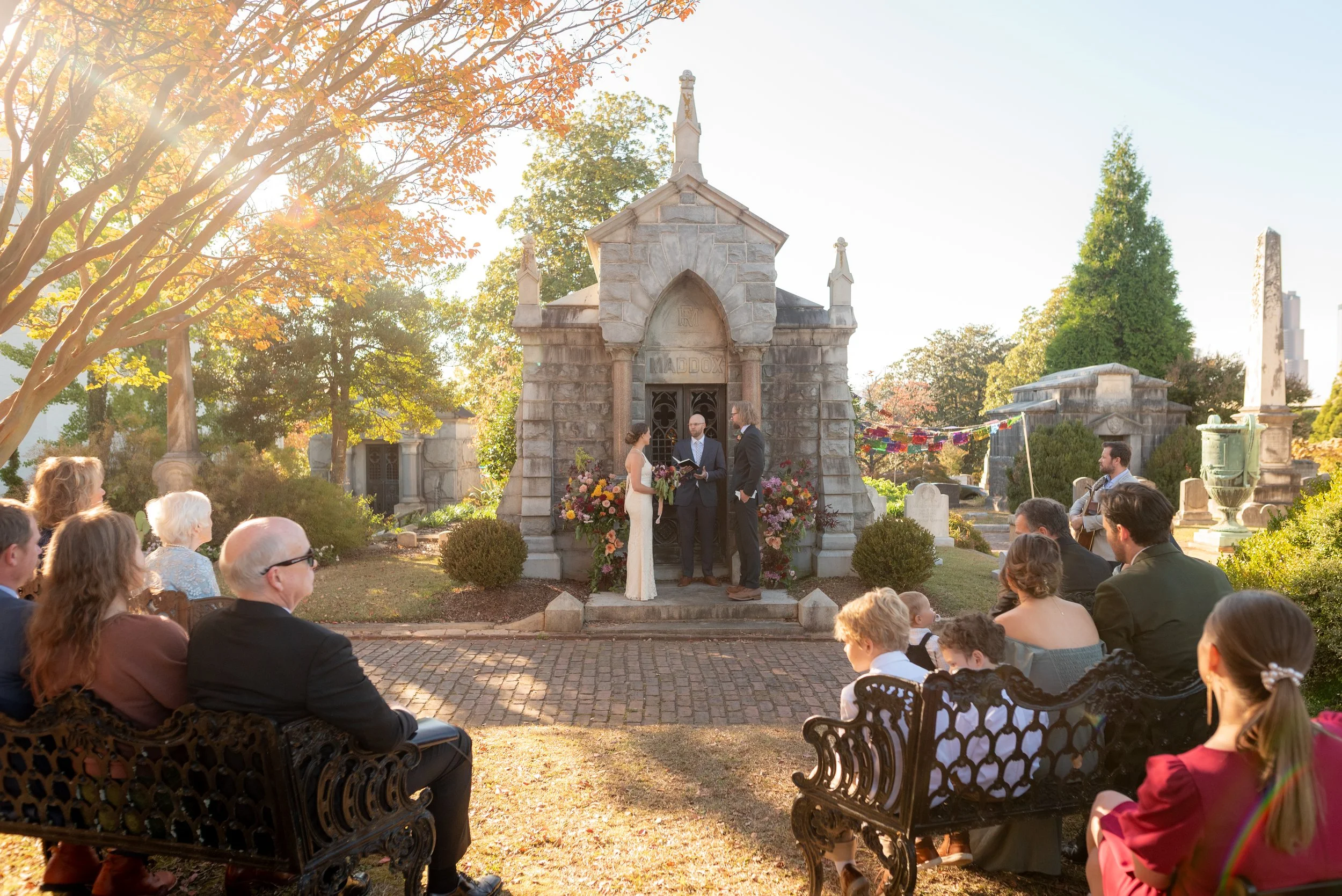 A wedding ceremony taking place in a cemetery, with a couple standing before an officiant, surrounded by seated guests. The ceremony is held at a small stone chapel or mausoleum, decorated with flowers. The scene is outdoors with sunlight filtering t