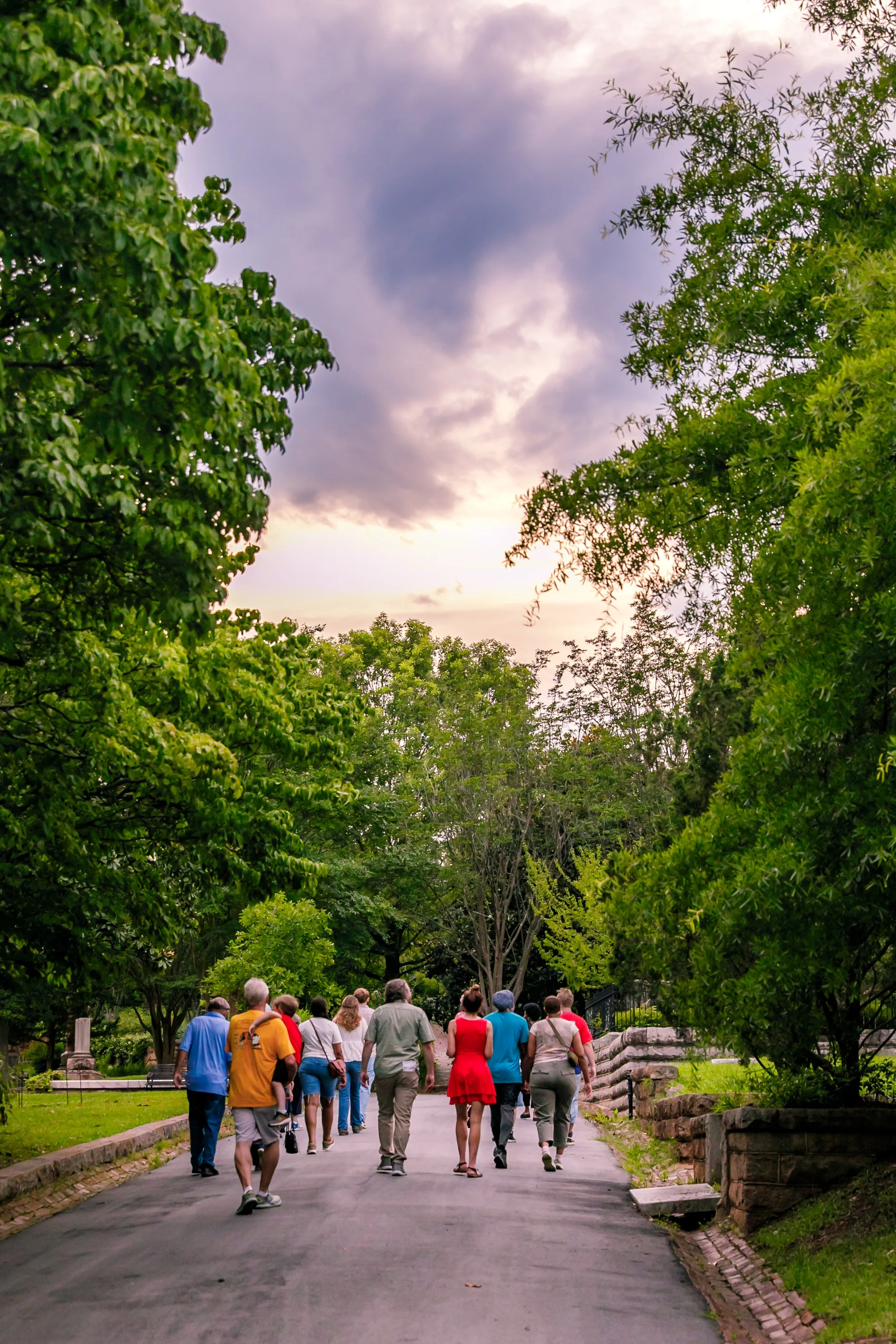 A group of people walking along a paved path in a park or garden, surrounded by green trees and foliage, with a cloudy sky overhead.