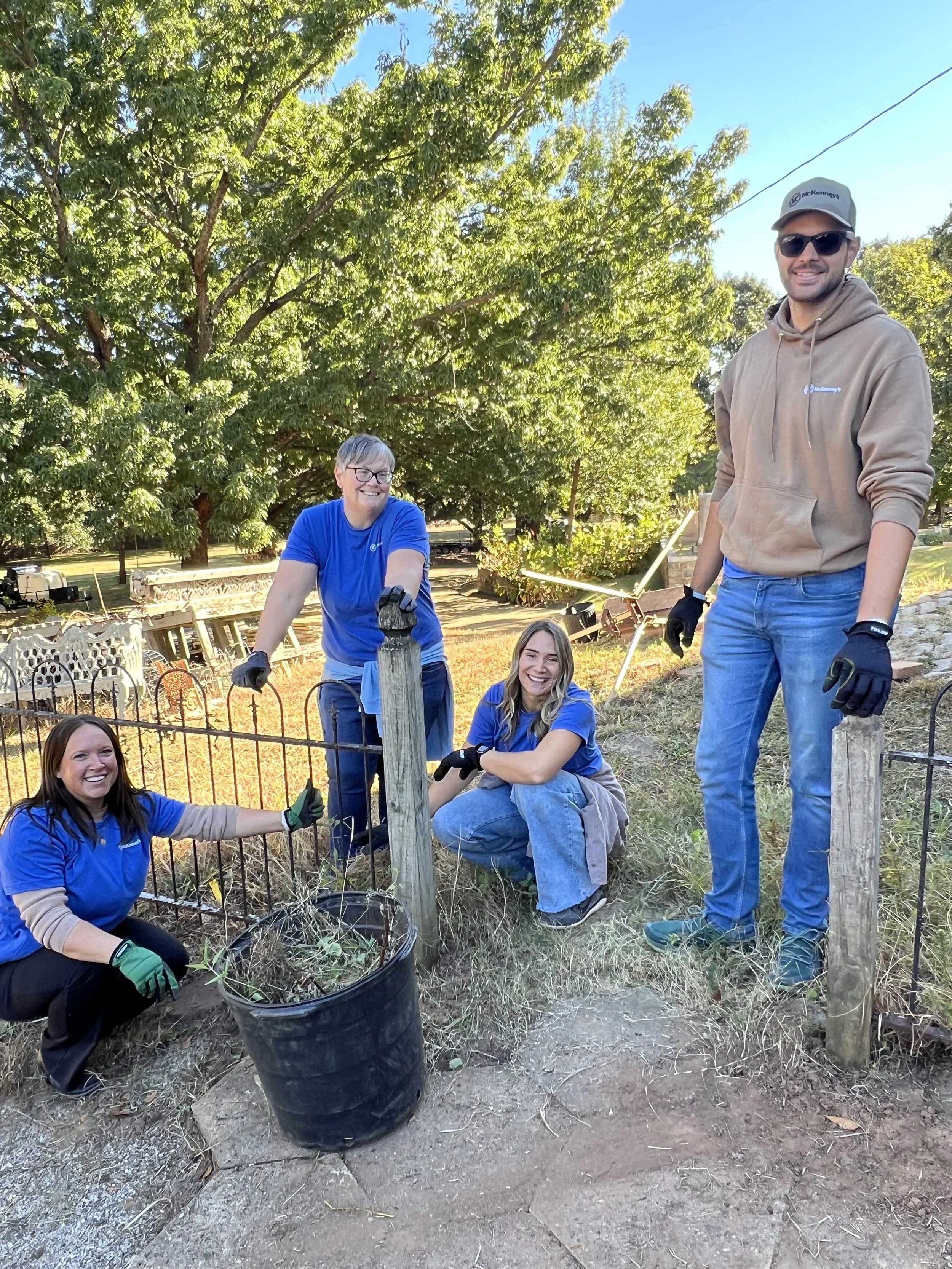 People working on a community gardening project outdoors, planting a young tree, all wearing gloves and casual clothes, smiling and surrounded by greenery.