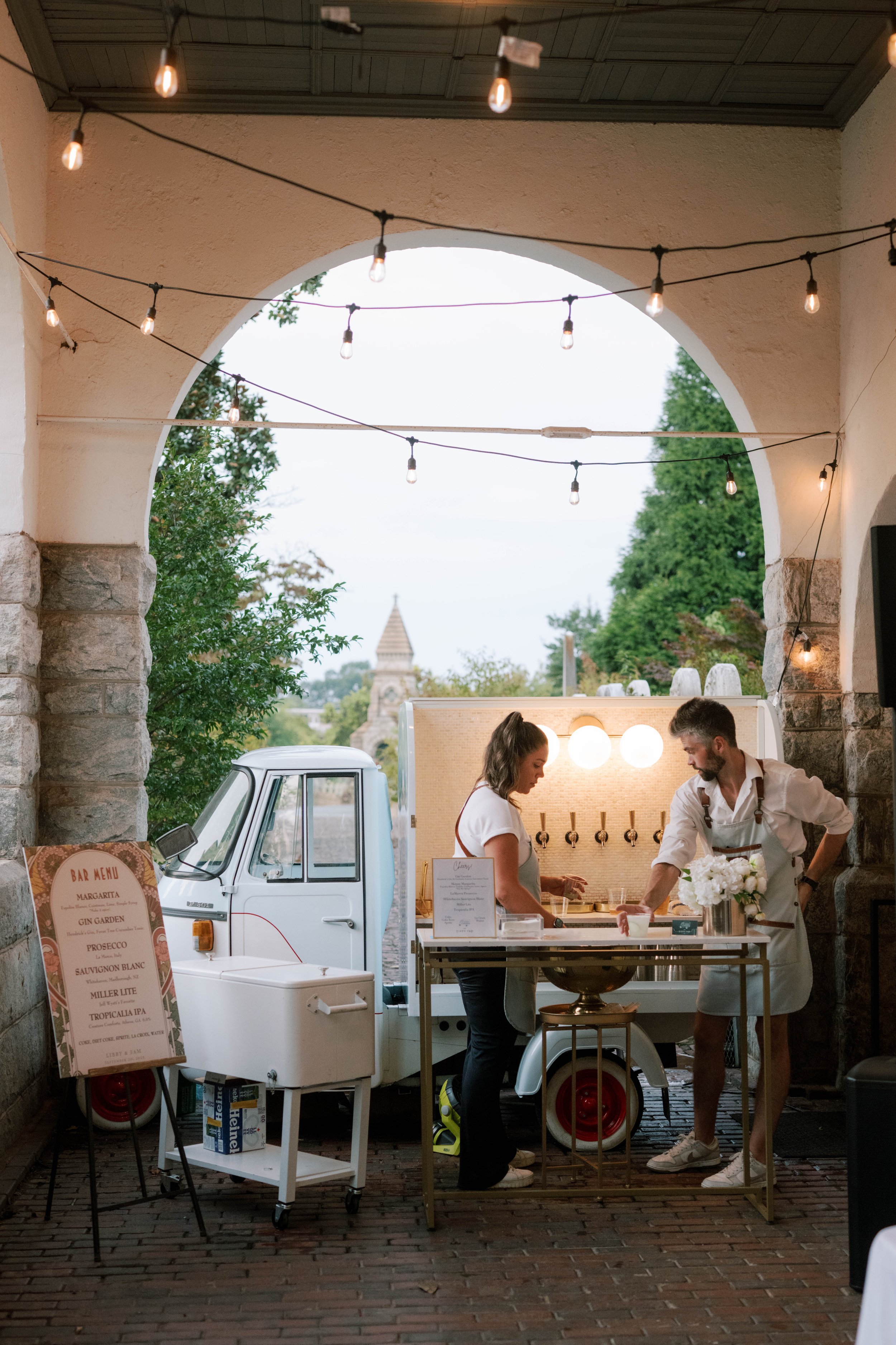 A mobile food stand in an outdoor covered area with string lights hanging above. Two people, a woman and a man, are working behind the stand, preparing food. A bar menu sign is visible on the left, and a white mini truck is parked behind the stand wi