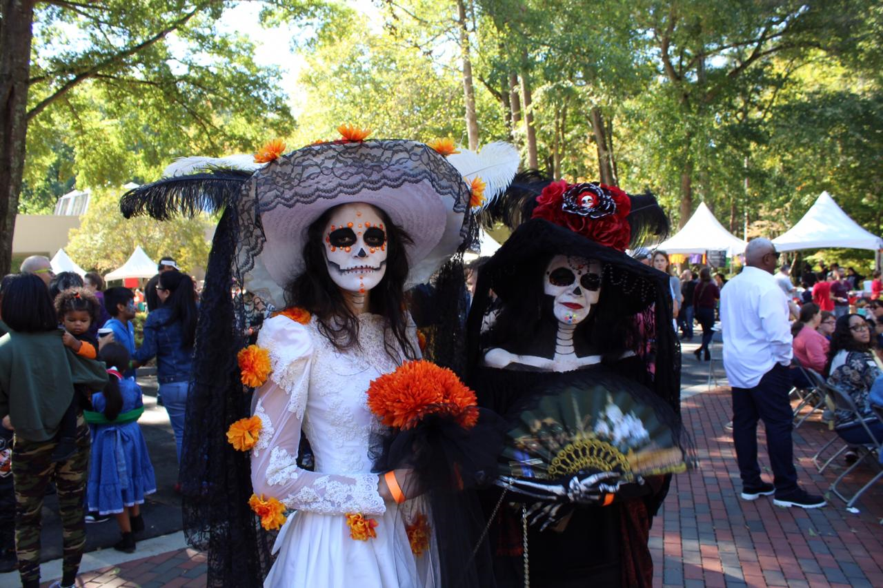 Two women dressed in Day of the Dead costumes, with painted skeleton faces, standing outdoors at a festival or celebration.