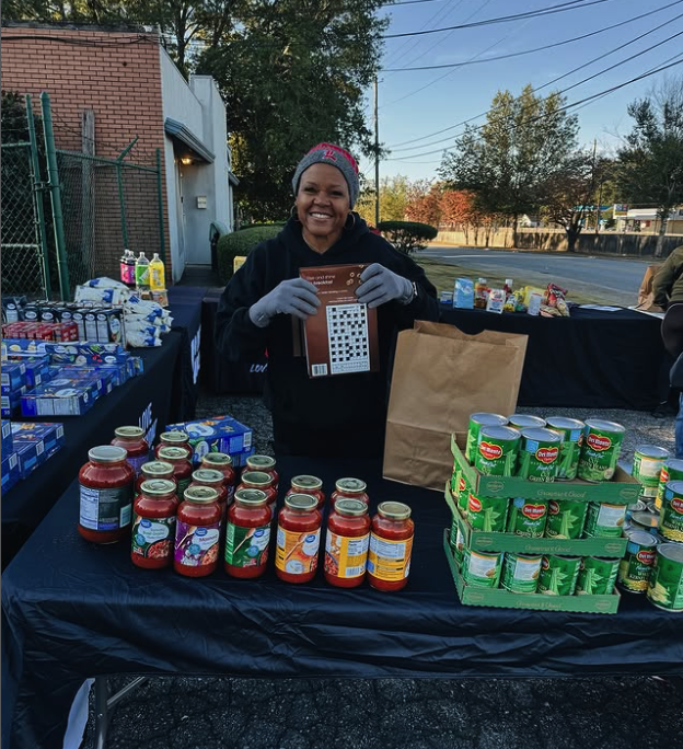 A woman smiling at a food drive table with canned goods and jars of sauce, holding a bingo card. Outdoors with trees and a road in the background.