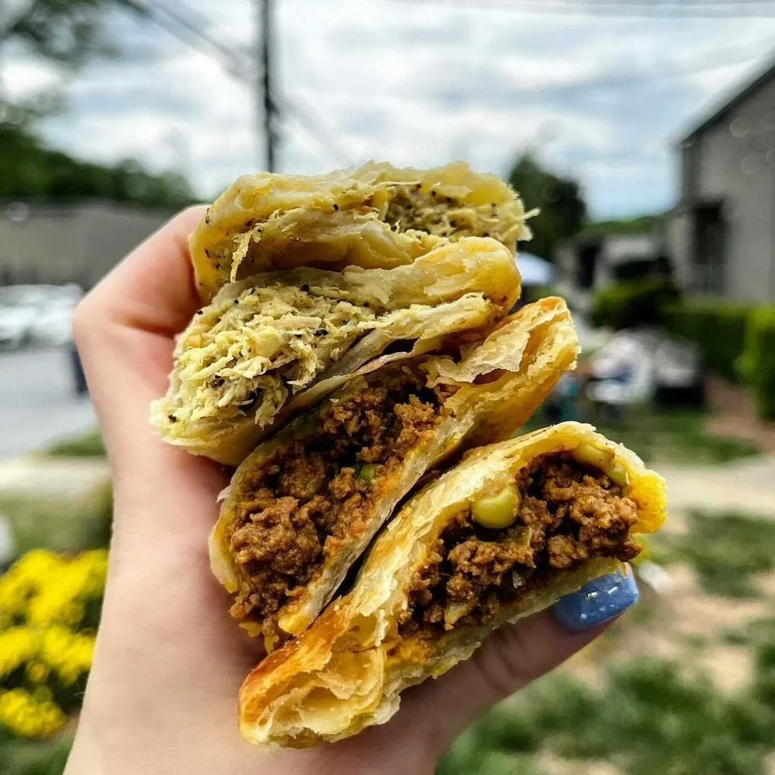 A hand holding three slices of meat-filled pastry, showing layers of flaky crust and ground meat filling outdoors with blurred background of houses and trees.