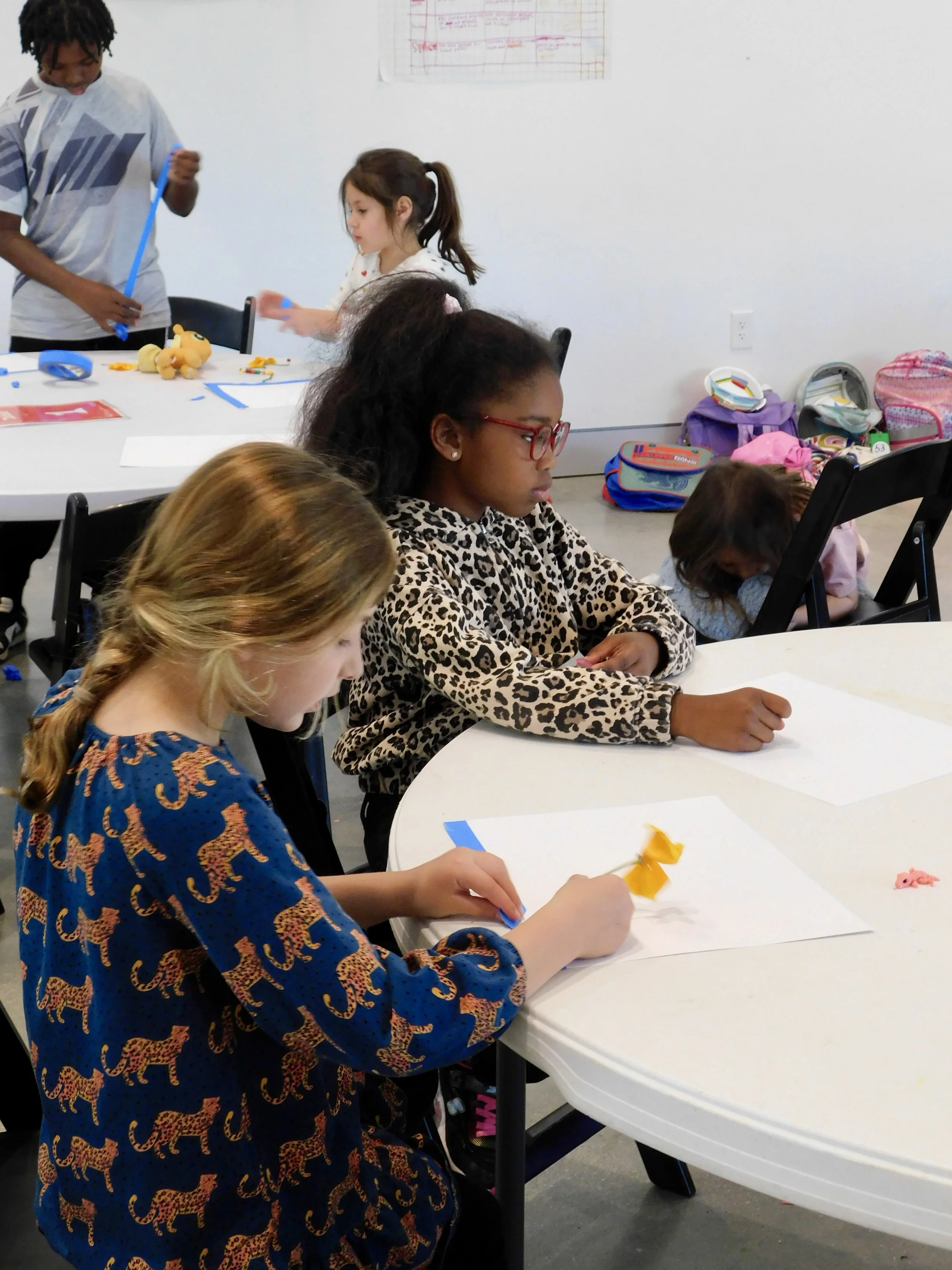 Children sitting at a white table engaging in arts and crafts, with backpacks and supplies on the floor, a boy and girl standing nearby.