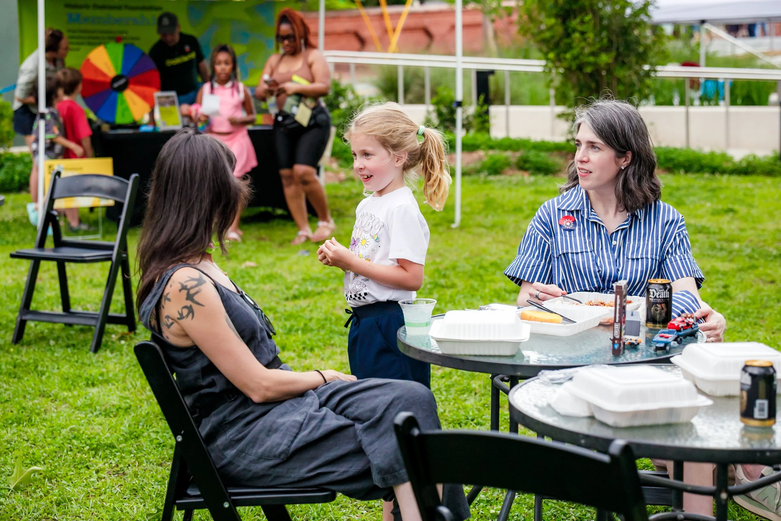 A group of people at an outdoor gathering, with a woman sitting in a wheelchair in the foreground, talking to a young girl. Other people are standing and sitting around tables with food and drinks. A tent with a colorful wheel and a food stand can be seen in the background.