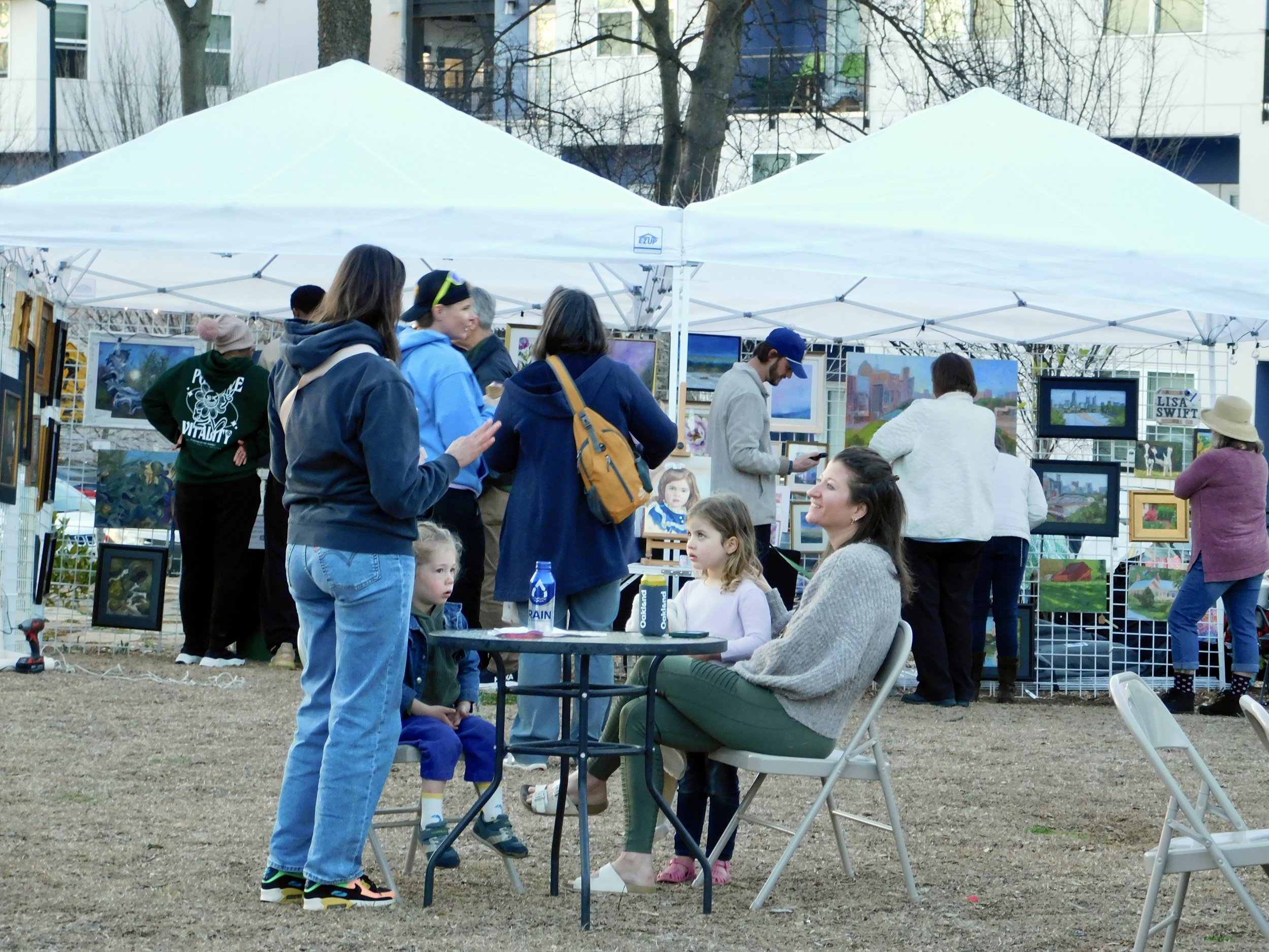 People at an outdoor art market, browsing paintings displayed under white tents, with some sitting at a table, and a variety of artwork including landscapes and portraits visible.