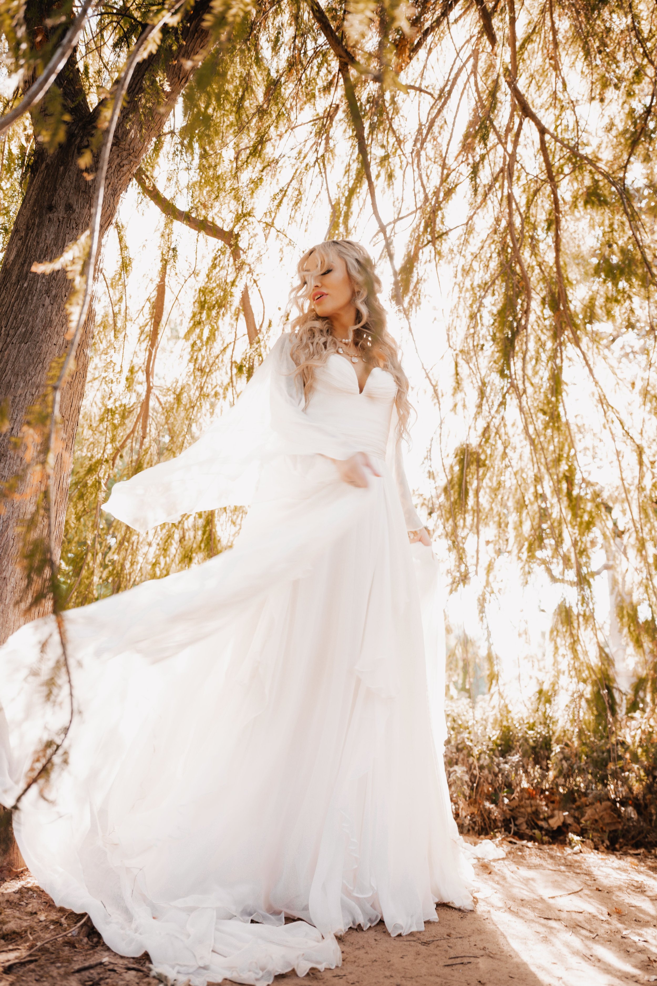 A woman in a flowing white dress stands outdoors among trees with sunlight filtering through the leaves.