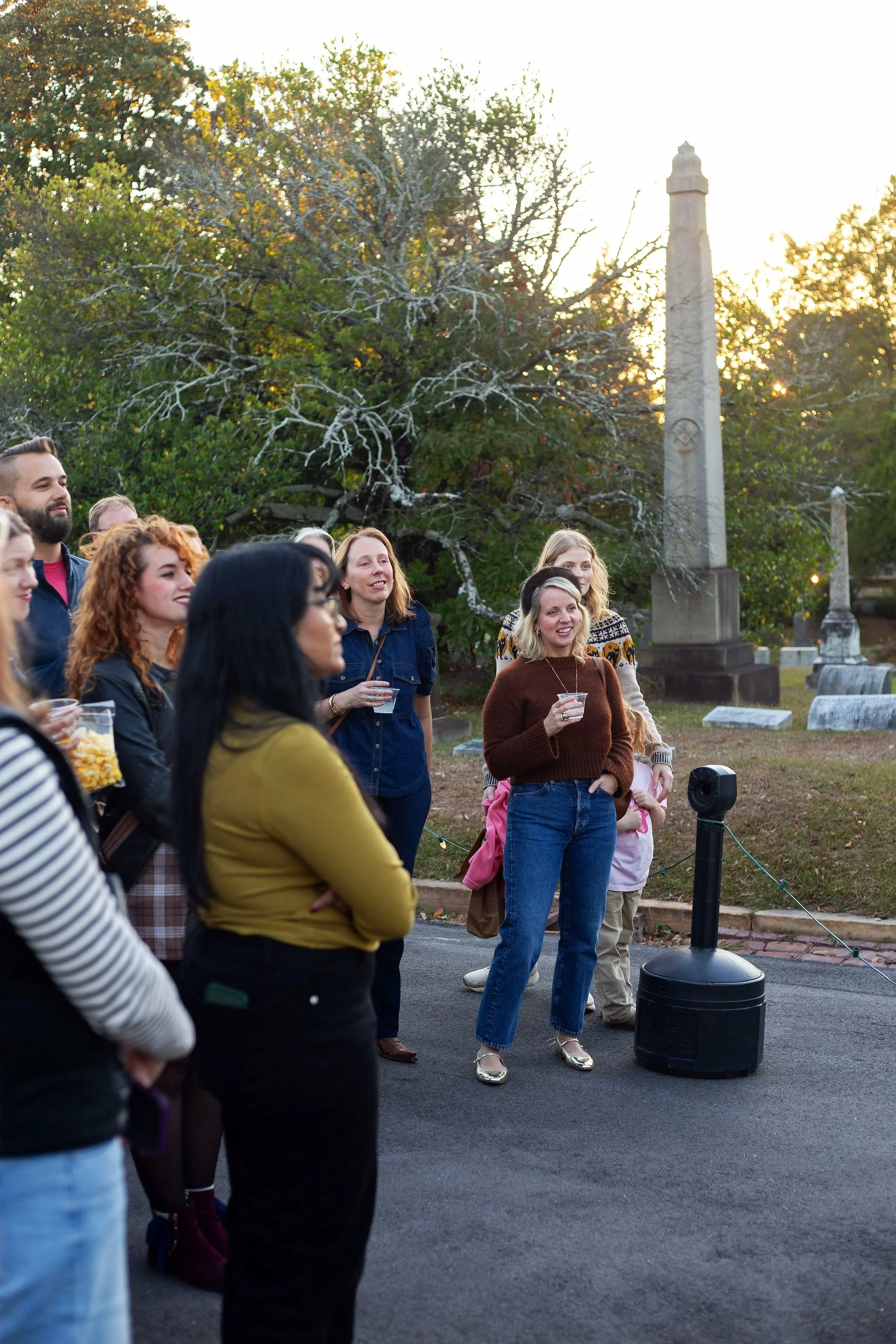 A group of people gathered outdoors during sunset, listening to a woman in a brown sweater and blue jeans speak. There are trees and a memorial stone in the background.
