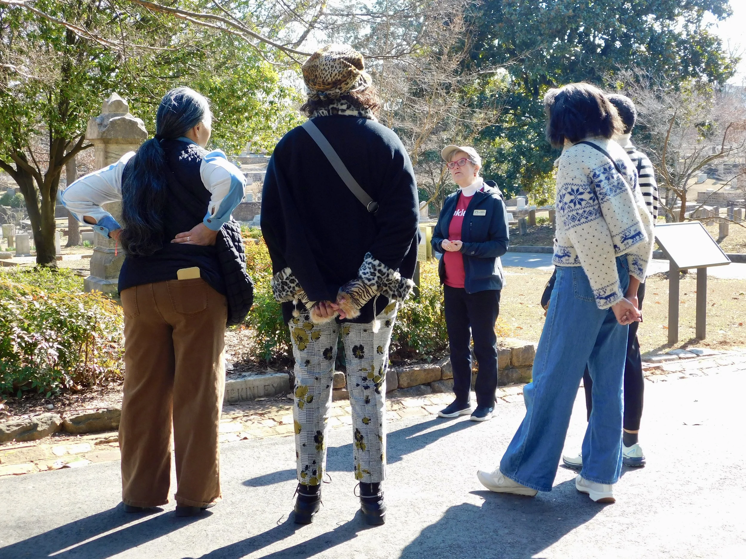 A group of five women standing outdoors in a park or garden, engaged in conversation. One woman, in the center, is wearing patterned pants and a leopard-print hat. The others are dressed casually, with some wearing jackets and sweaters. There are trees and shrubs in the background, and a stone marker in the ground.