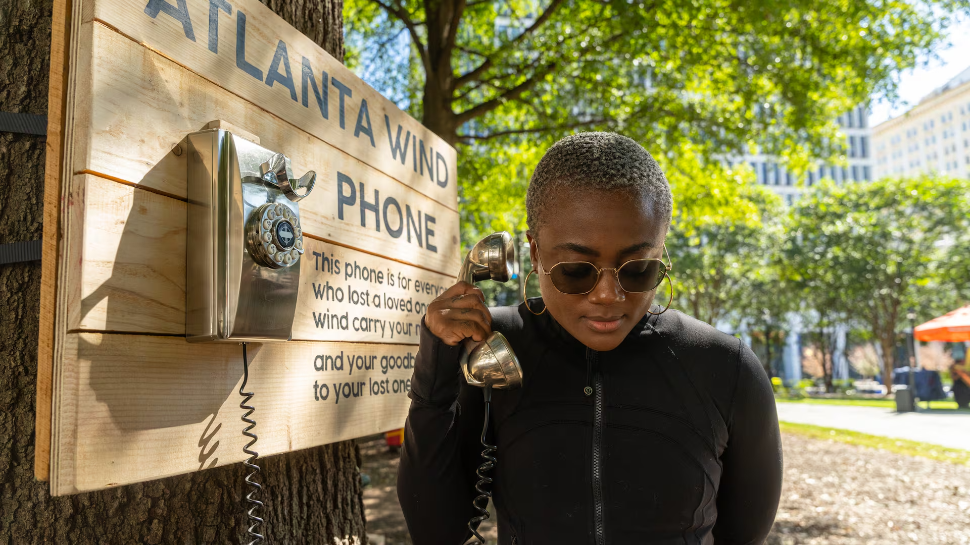 A woman with short hair and sunglasses talking on a vintage payphone mounted on a tree at a park. There is a wooden sign with the words 'Atlantic Wind Phone' on it, attached to the tree. Sunlight filters through green leaves in the background.