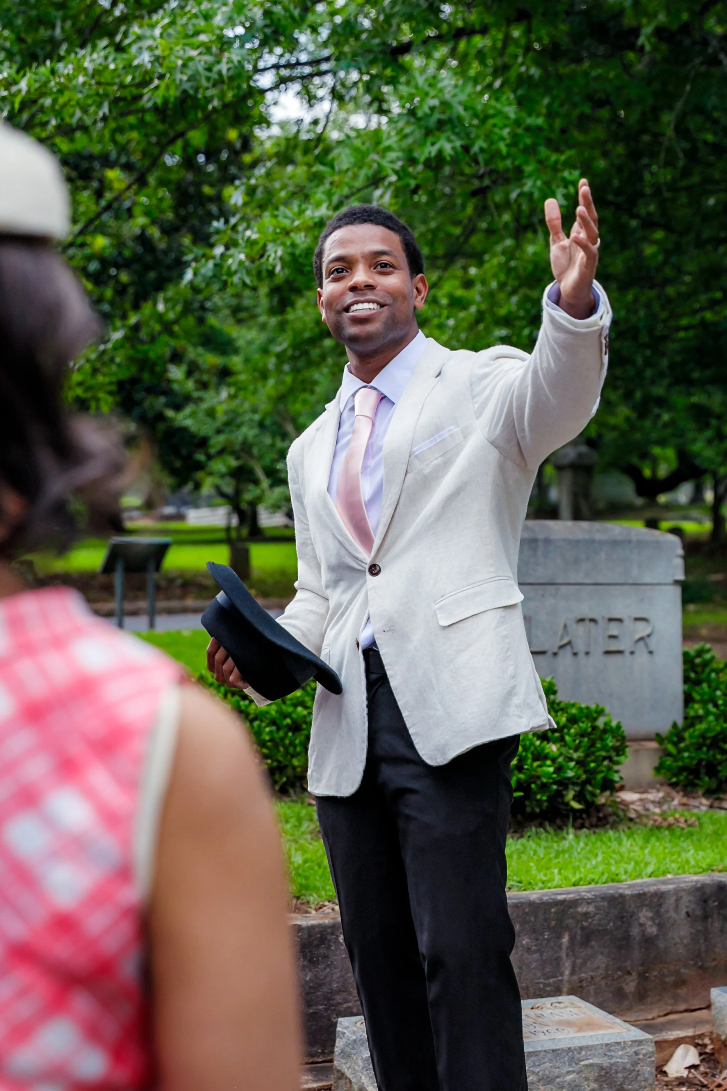A man in a light-colored suit and tie is smiling and speaking to a person in a pink dress and a white hat, with a woman in the foreground. They are outdoors near a cemetery with trees and gravestones.