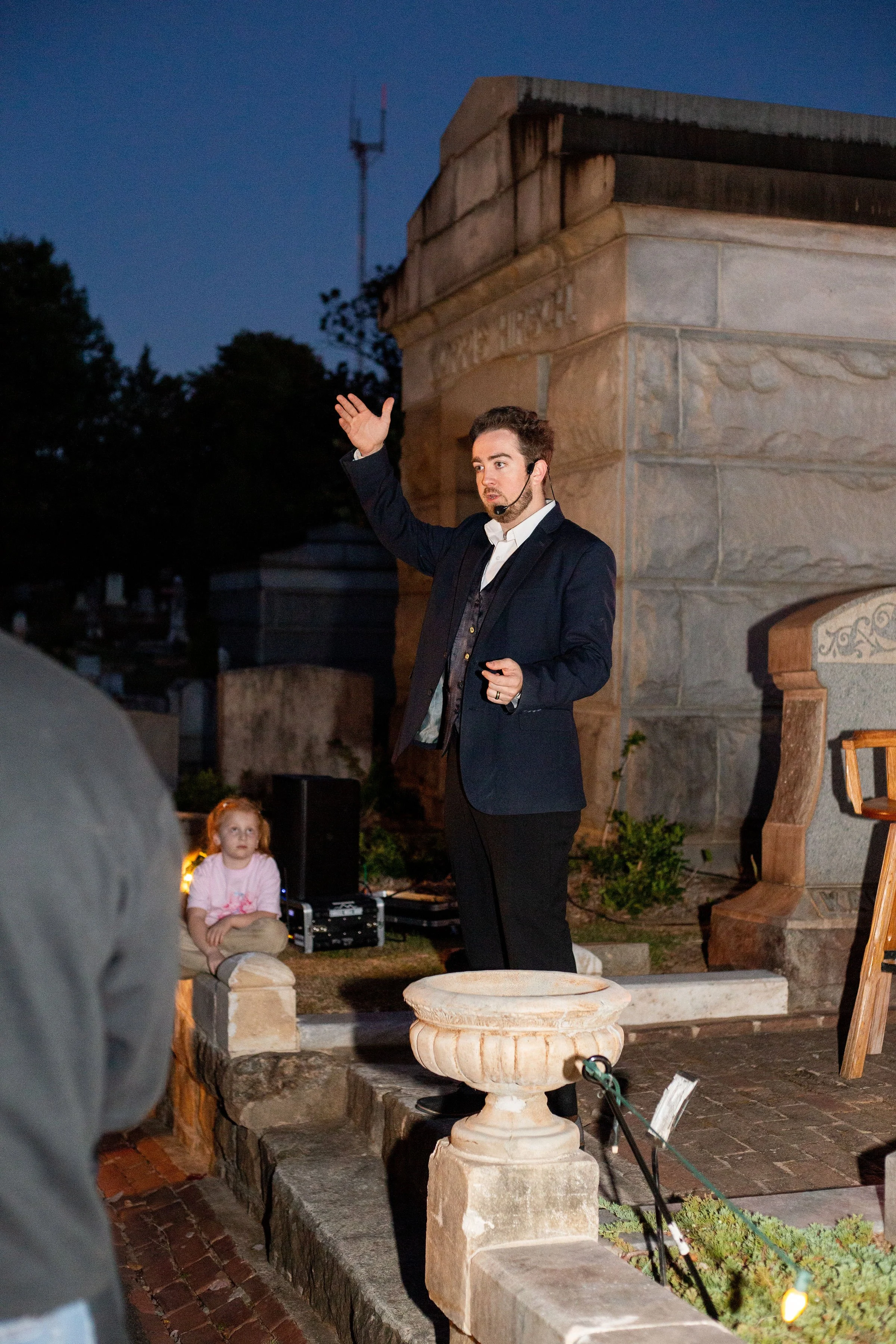 A man in a suit speaking to an audience at night, with a young girl sitting on a stone ledge nearby. The background features a stone monument and a tall antenna, with trees and a dark blue sky.