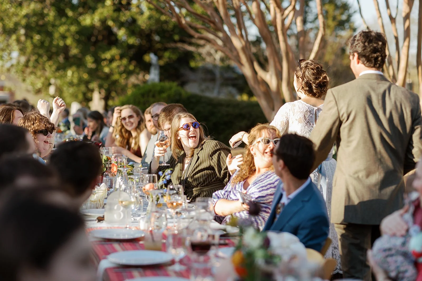 People sitting at outdoor banquet table, laughing and socializing, with two standing individuals engaging with the seated guests, surrounded by trees and sunlight.