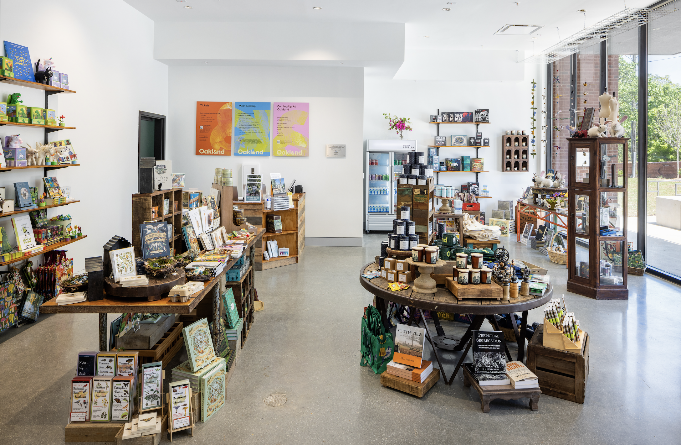 Interior of a gift shop with shelves and tables displaying books, candles, toys, and souvenirs, with large windows on the right letting in natural light.