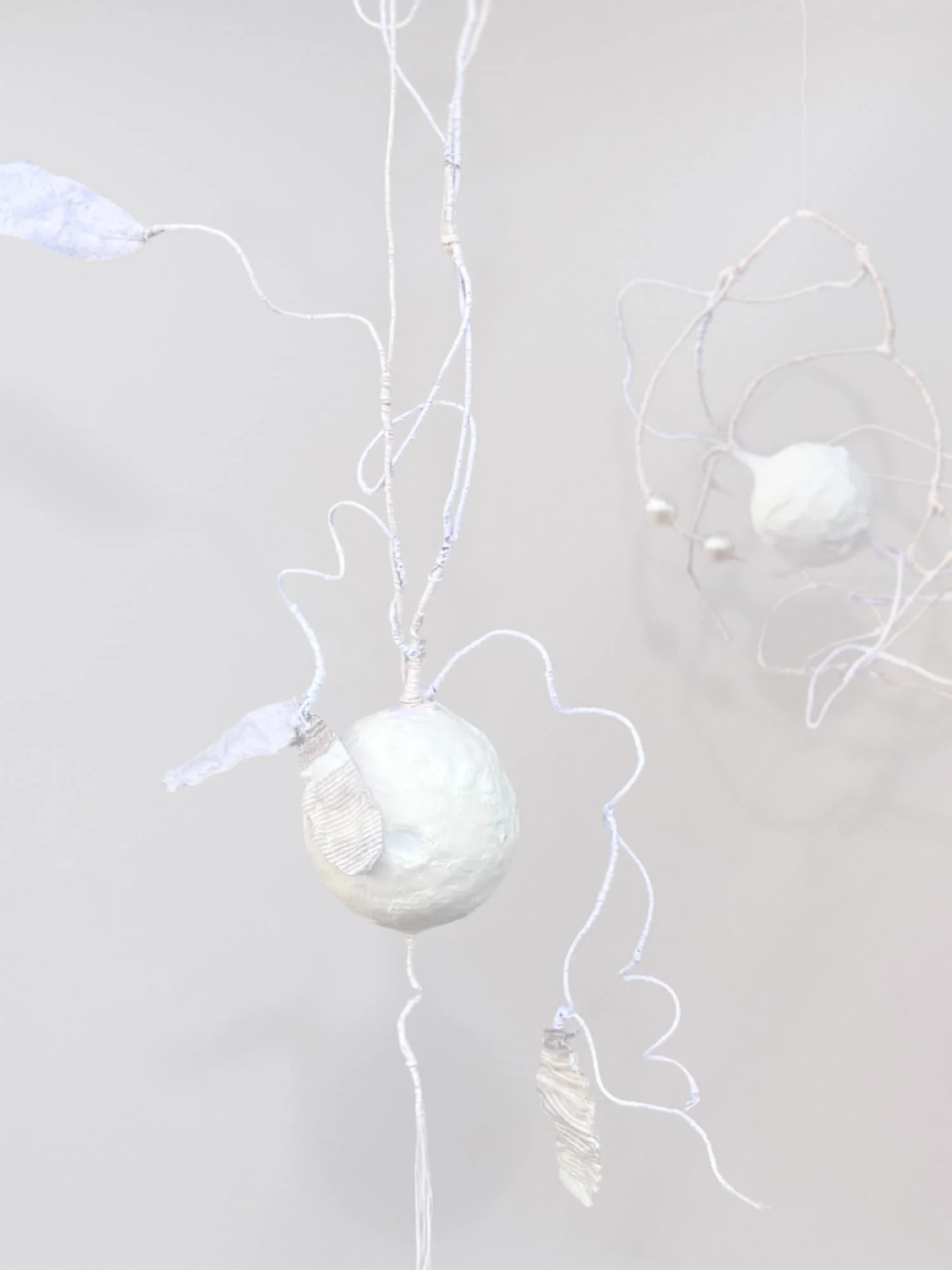 White sculptural orb suspended against a pale gray background with long curling threads and root-like strands extending upward and outward in irregular organic lines.
