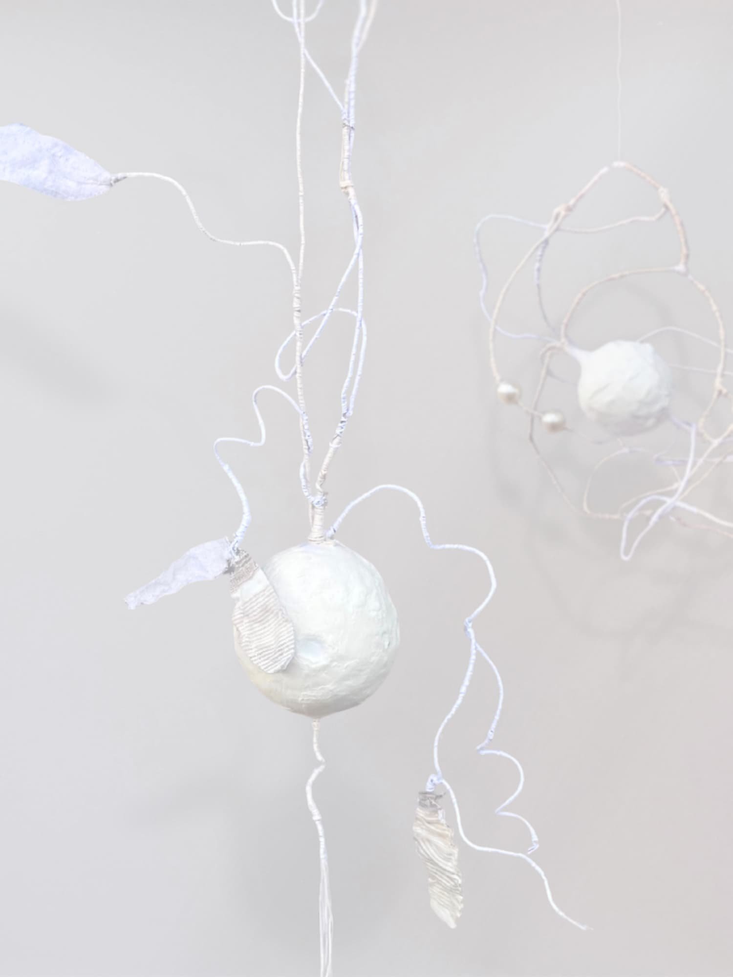 White sculptural orb suspended against a pale gray background with long curling threads and root-like strands extending upward and outward in irregular organic lines.