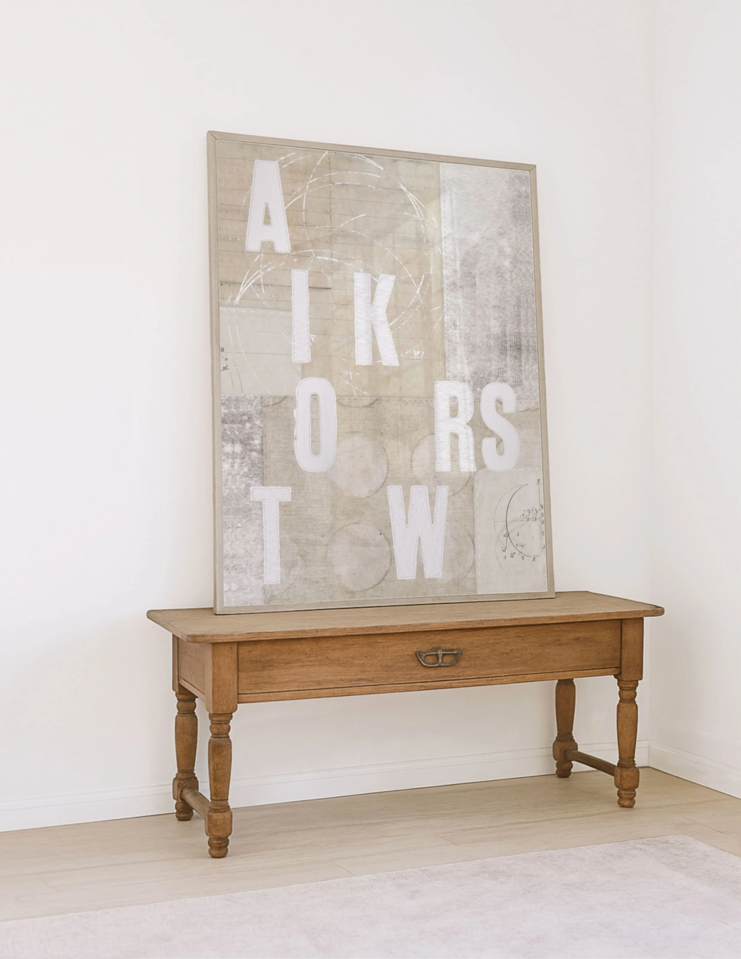 A wooden table with turned legs and a driftwood framed textile art piece resting against the white wall behind it.
