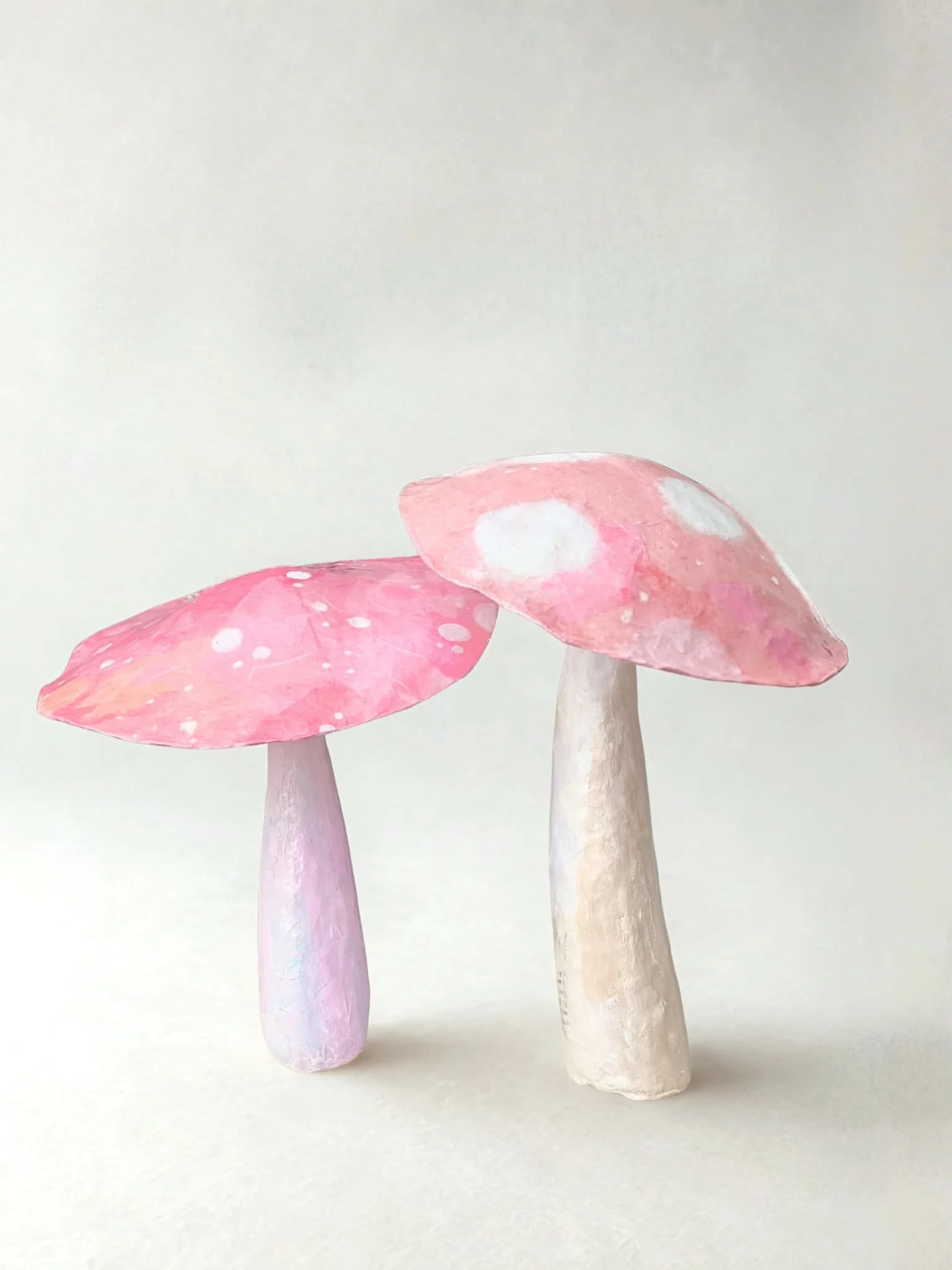 Three paper mache mushrooms with pink and white caps and white stems, set against a plain white background.