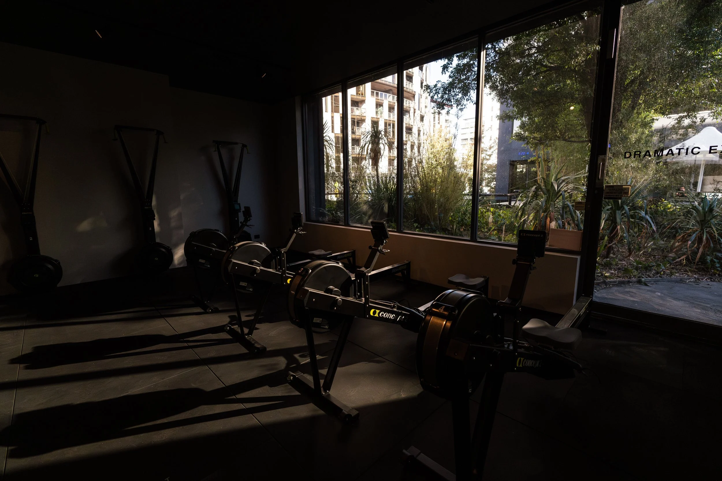 Indoor fitness room with rowing machines, dimly lit, large windows showing outside greenery and buildings.