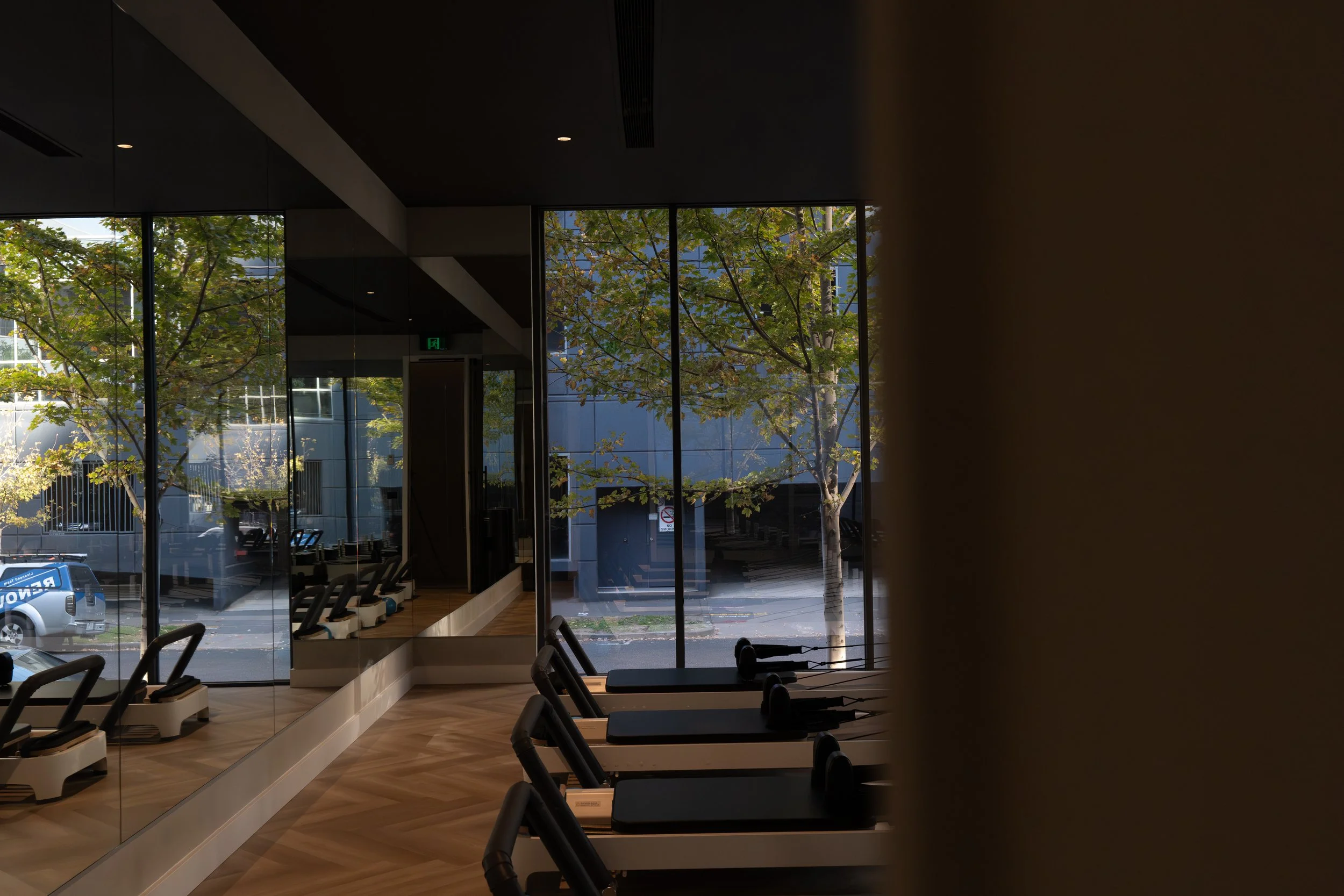Empty gym with treadmills facing large windows showing trees and a street outside.