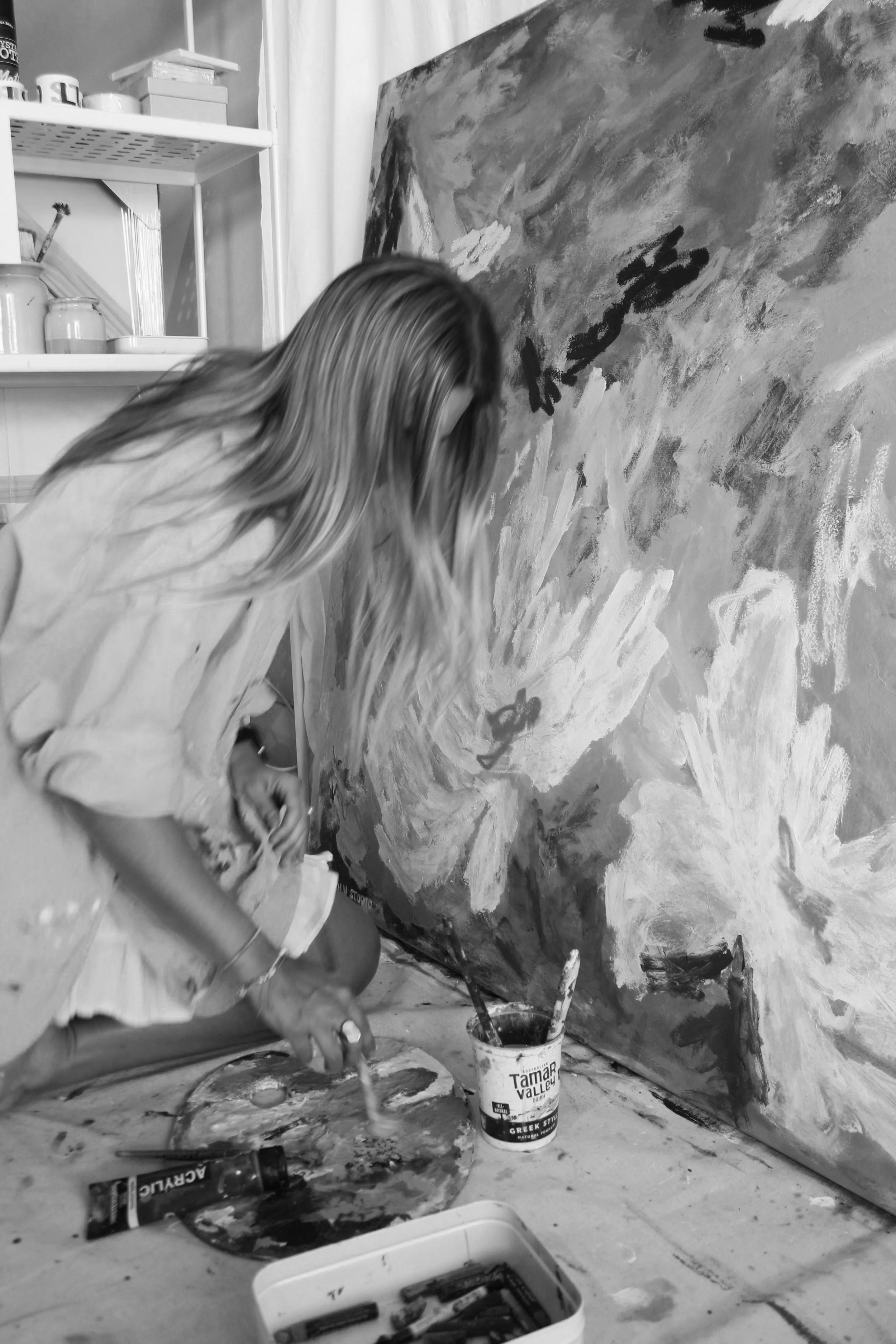 A woman with long hair painting an abstract mural on a large canvas in a studio, surrounded by art supplies.