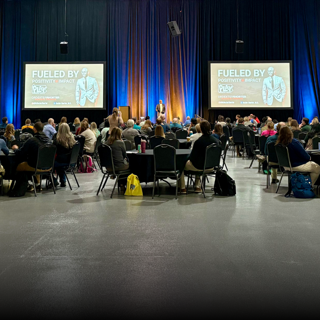 This is Dr. Dexter Shorter delivering a keynote speech at a conference room filled with attendees facing a stage where a speaker is presenting. Large screens display the title 'Fueled by Positivity Impact' and the name Dr. Dexter Shorter.