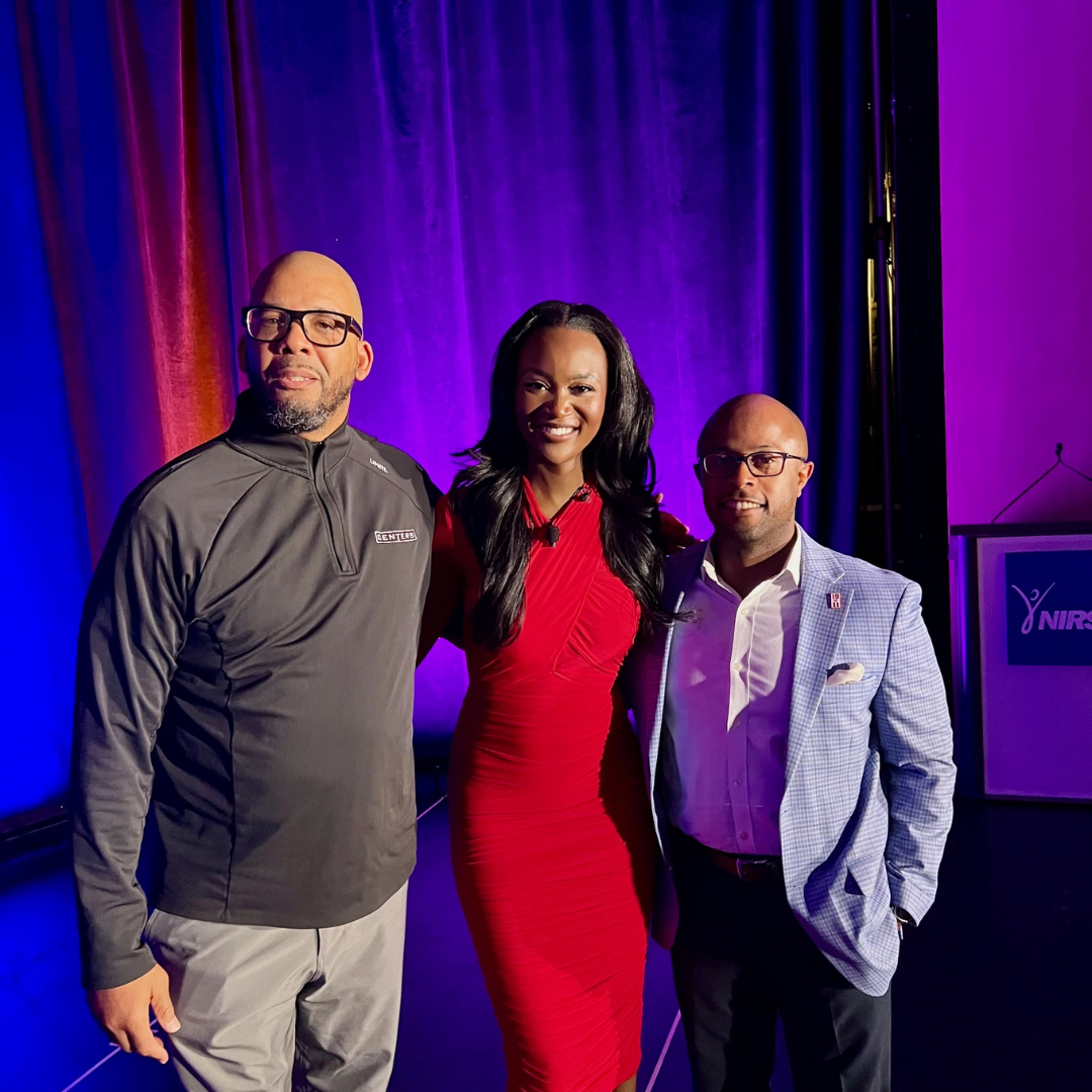 This is Dr. Dexter Shorter standing with two people on a stage with purple and red stage curtains in the background. The woman in the center is wearing a red dress, and the two men on either side are dressed in business casual attire.